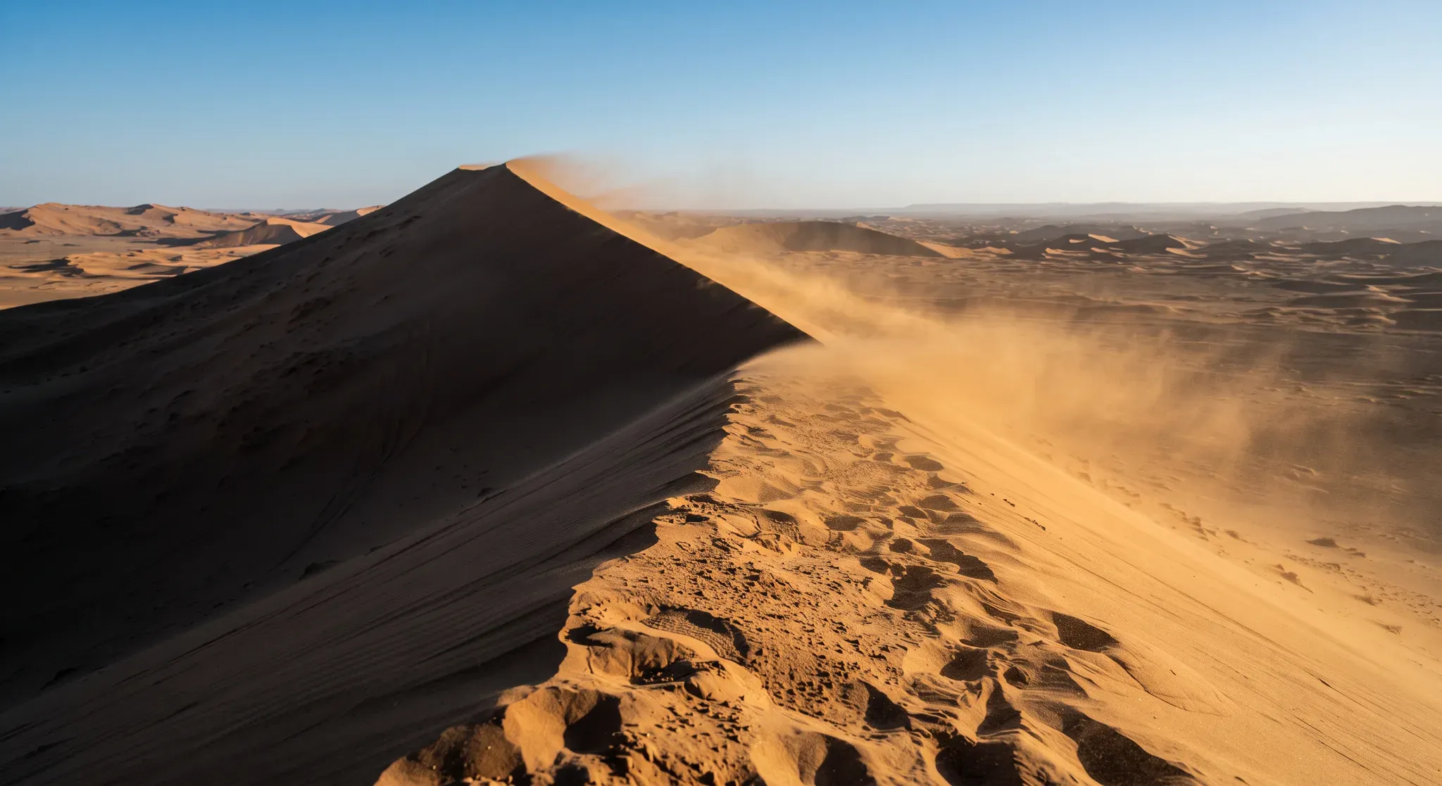 A dramatic high-angle view along a massive, sharply ridged sand dune in an expansive desert. Strong directional sunlight from the right illuminates the windward face and foreground sand in warm golden-orange tones, while the steep leeward face is cast in deep shadow of dark brown. Wind actively strips sand off the dune crest, creating a visible streaming dust plume that drifts leeward across the sunlit face. Footprints are visible in the textured foreground sand near the ridge base. A vast field of undulating dunes extends to the horizon under a clear deep-blue sky, with a thin dust haze softening the mid-distance. No people or animals are present.