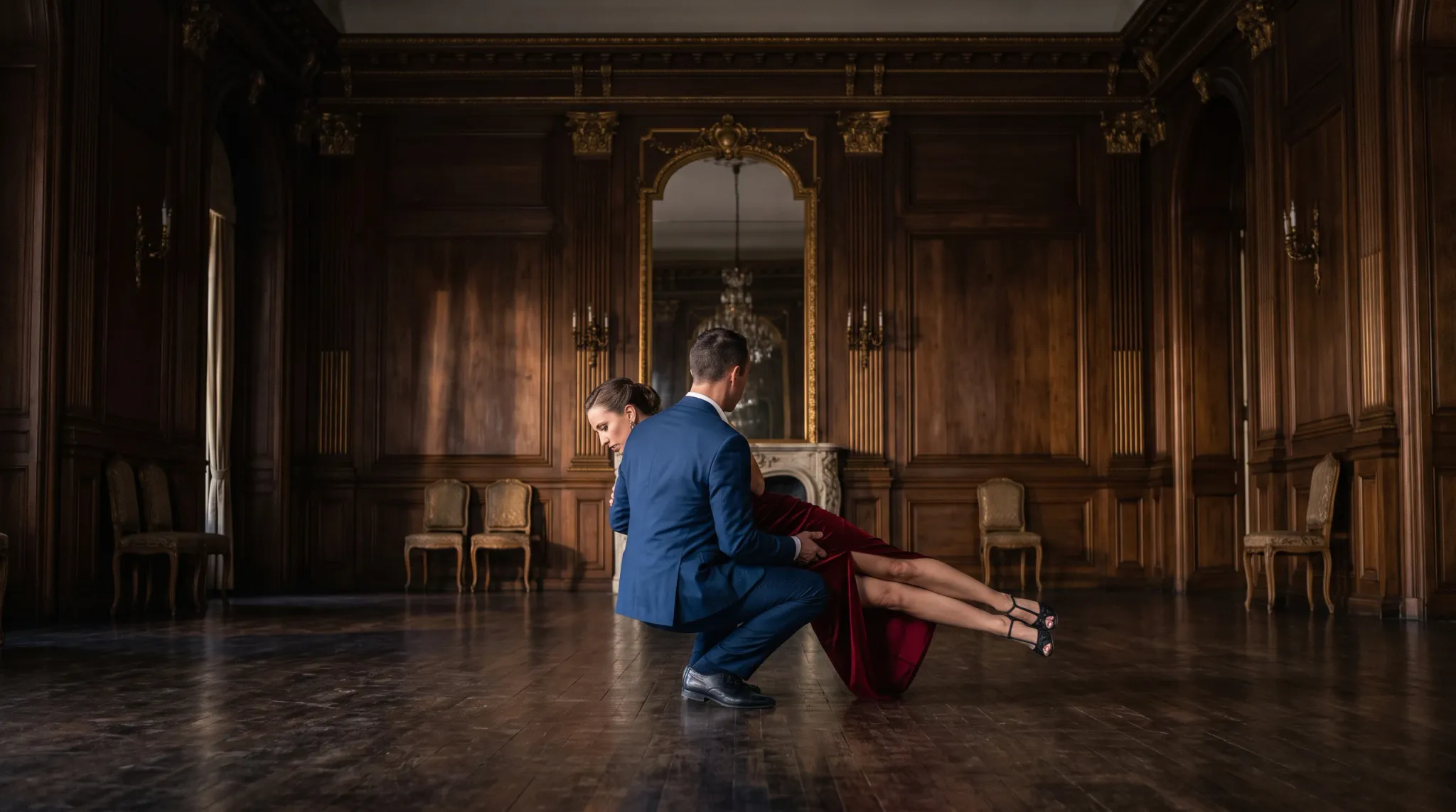 A man in a tailored blue suit and black dress shoes lowers into a deep crouch to support and carry a woman in a dramatic horizontal ballroom dance dip. The woman wears a floor-length red evening gown with a high slit and black strappy heels. They perform in the center of a spacious, opulent ballroom featuring rich dark wood wall paneling, Corinthian pilasters, gilded accents, and an arched mirror over a fireplace mantle.