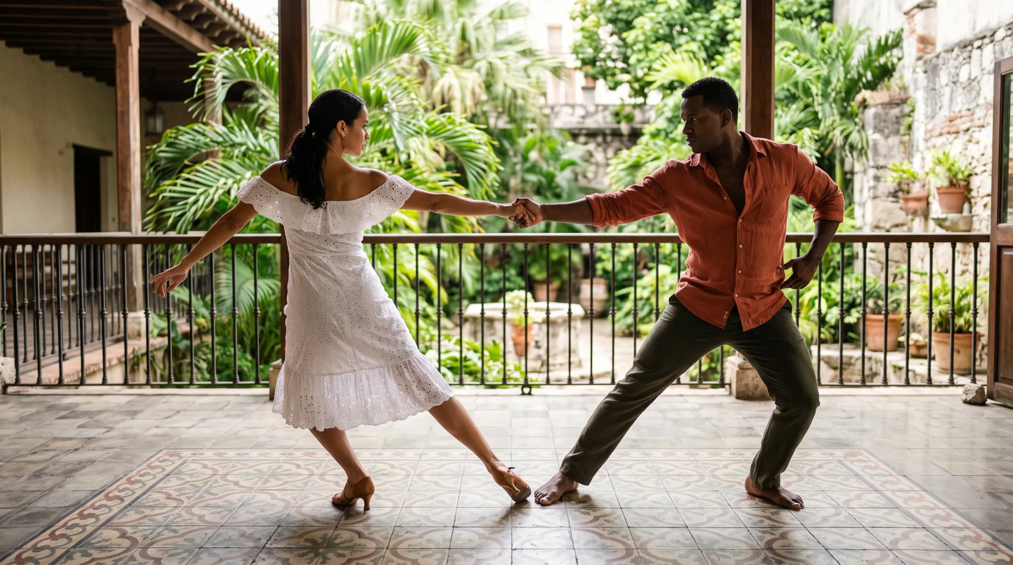 Two adults dance together on a covered tropical patio, holding hands in an open partnered pose. The woman wears a white off-shoulder dress and brown heeled shoes; the man wears an orange long-sleeve shirt and dark trousers and is barefoot. Lush palms, tiled flooring, railings, and stone walls form the courtyard setting.