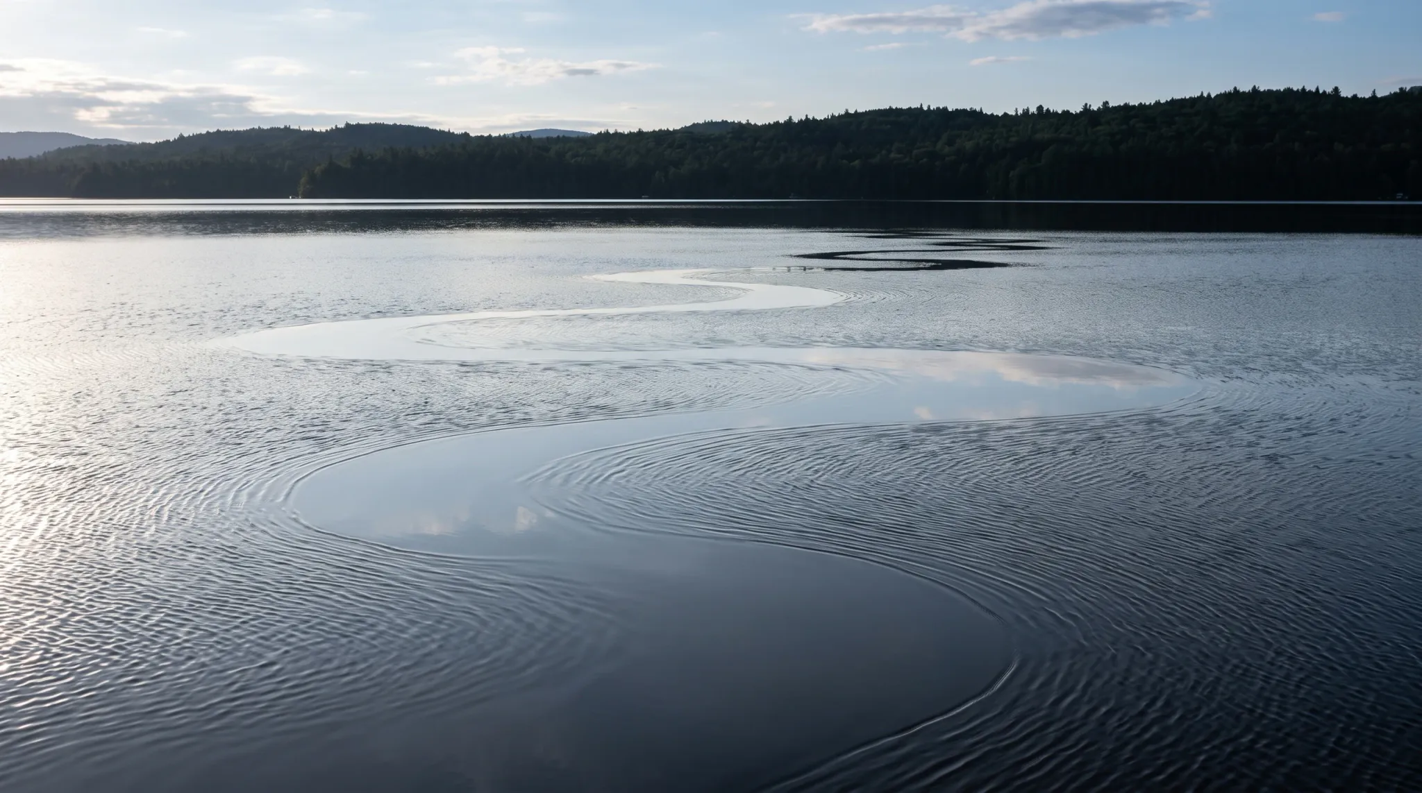 A tranquil wide-angle view across a calm wilderness lake, where a winding serpentine band of glassy water creates an S-curve through concentric surface ripples, reflecting the pale morning sky against a backdrop of forested hills.