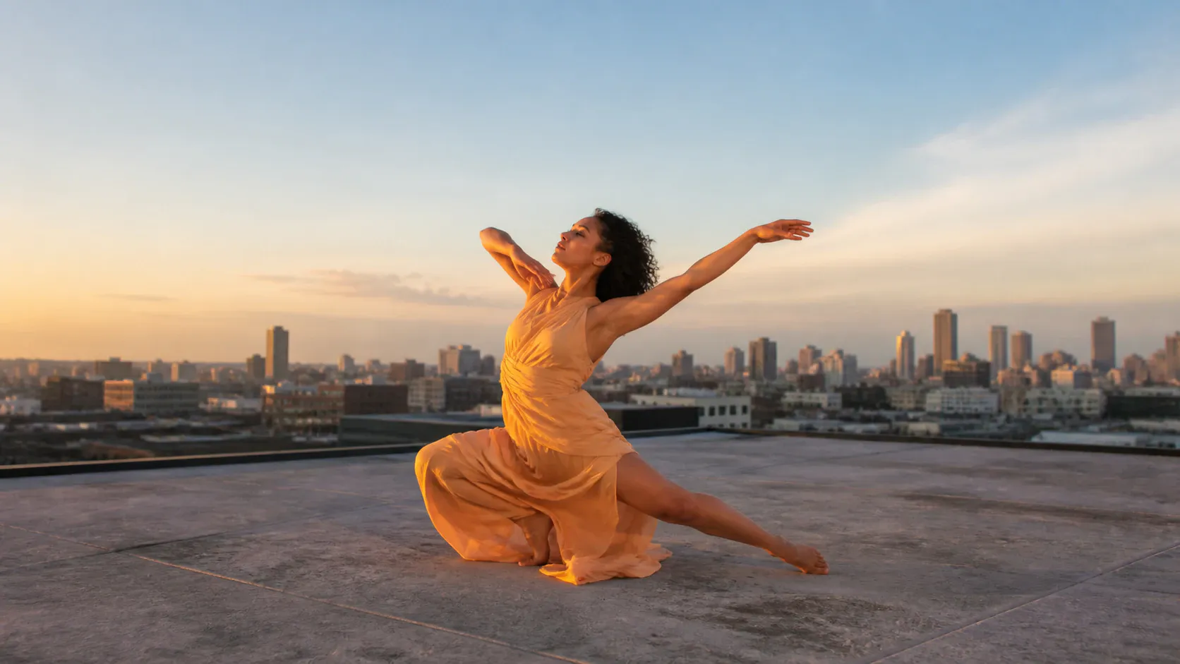 A solo dancer in a flowing orange dress holds an expressive kneeling pose on a concrete rooftop, with arms extended and a city skyline behind her under a broad sunset sky.