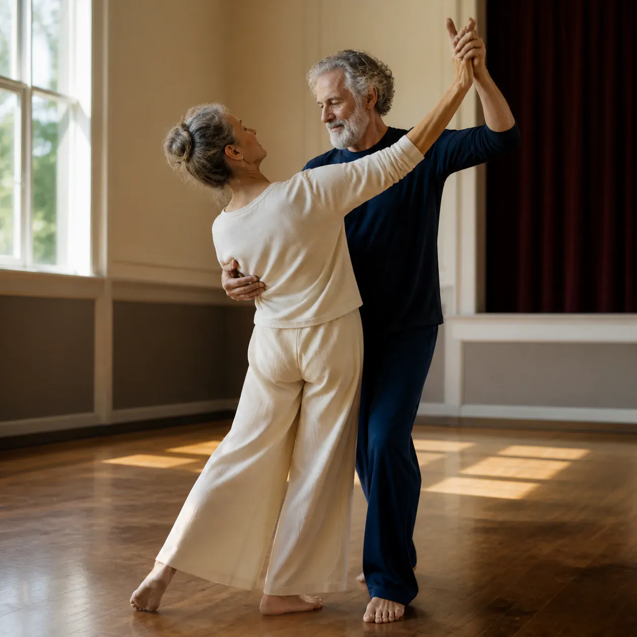 An older man with gray hair and a full beard dances with an older woman whose hair is tied in a high bun. Both dancers are barefoot on a polished hardwood floor inside a bright studio with natural window light. The woman is dressed in a cream long-sleeved top with wide-leg trousers, and the man wears a navy blue long-sleeved shirt and trousers. They hold hands in a partner dance pose with gentle, engaged expressions.