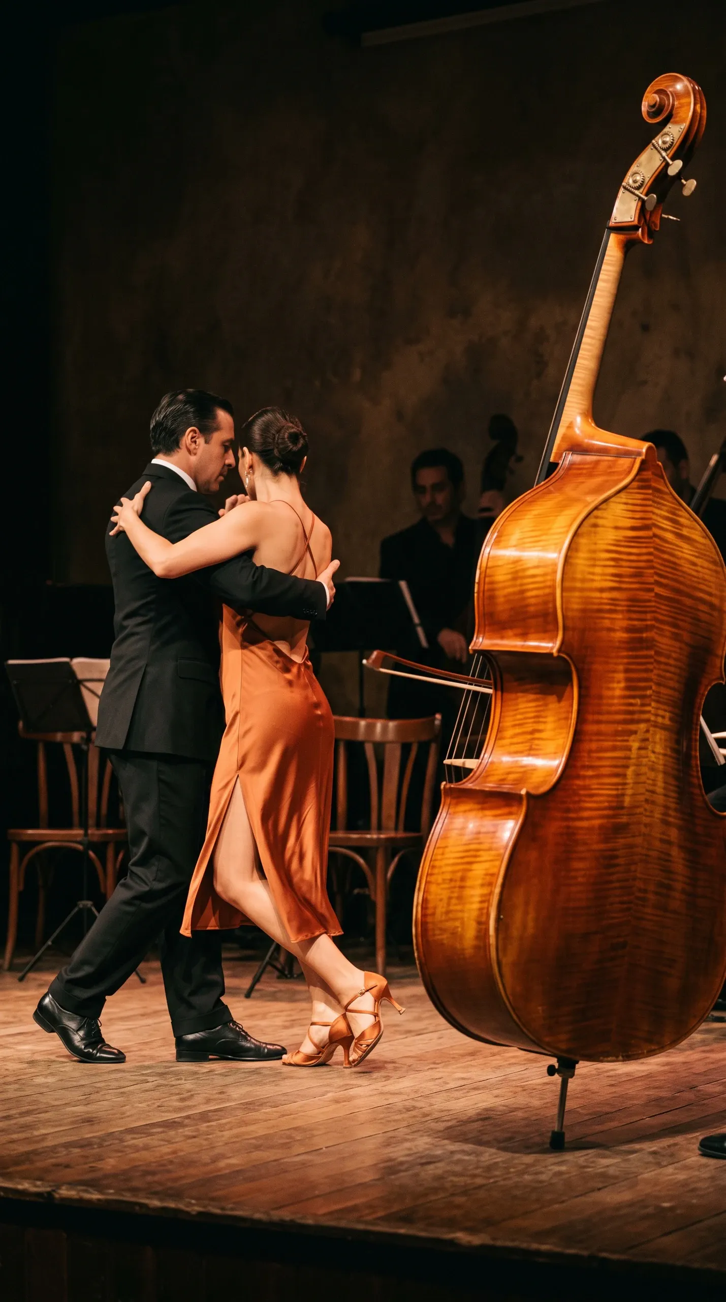 A formally dressed couple performs a close tango-like dance on a wooden stage beside a large polished double bass. Warm theatrical lighting illuminates the dancers and instrument while several partially obscured musicians and music stands recede into the dark concert venue.
