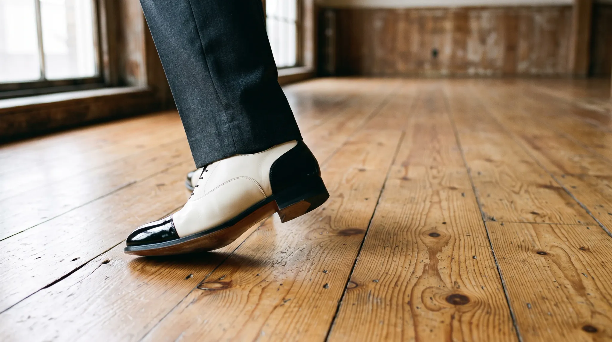 A low-angle shot capturing the lower legs of a person walking forward on a wide-plank wooden floor. The person is dressed in dark gray trousers and a distinctive pair of black and white leather spectator dress shoes. The room is indoors and appears relatively empty, illuminated by natural daylight coming from large windows in the background.