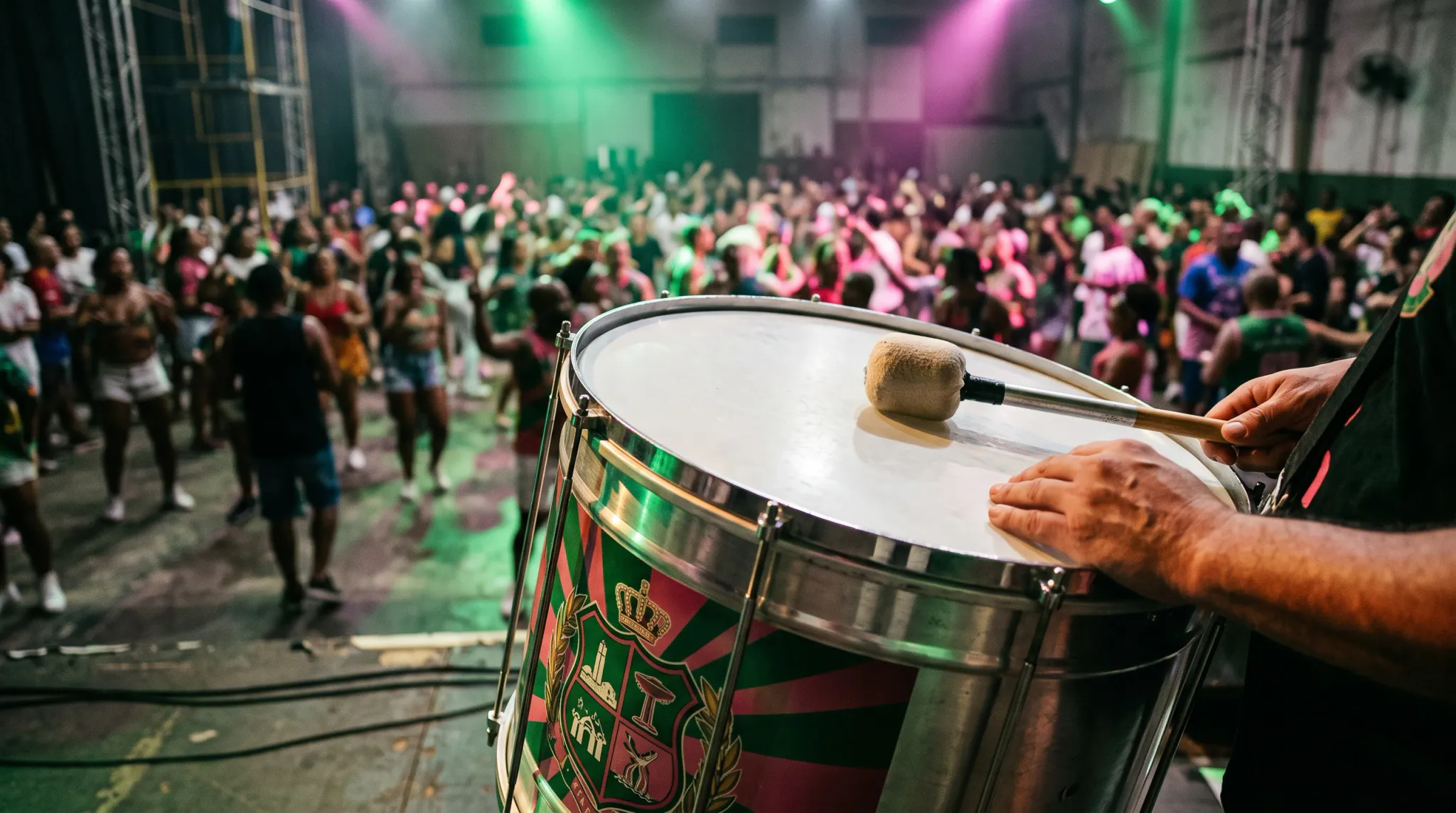 A cropped drummer in a black garment plays a large red, green, gold, and silver decorated drum with mallets in the foreground while a large crowd dances in a brightly lit indoor concert or dance venue.
