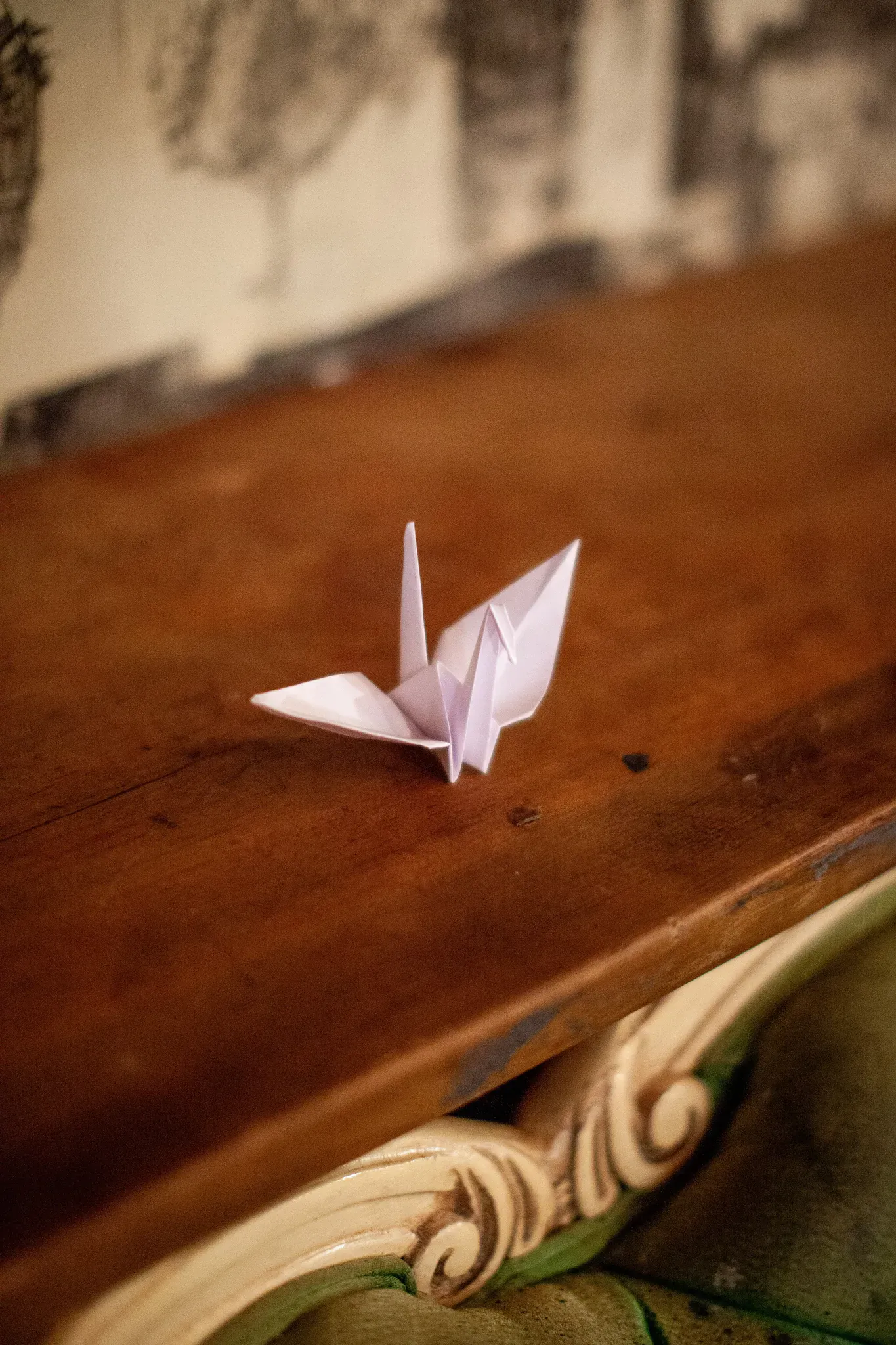 A small white origami paper crane rests on a warm brown wooden tabletop. The crane is sharply emphasized against a softly blurred indoor background with ornate carved furniture and wall artwork. The portrait photograph uses shallow depth of field and warm, diffuse lighting.