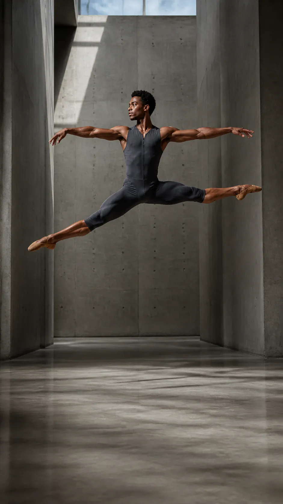 A single male-presenting ballet dancer in a black sleeveless skin-tight unitard and beige pointe shoes performs a suspended split leap inside a tall, minimalist concrete interior. His arms extend horizontally, his face turns left, and strong sunlight enters from high windows, casting angular shadows across the gray walls and polished floor.