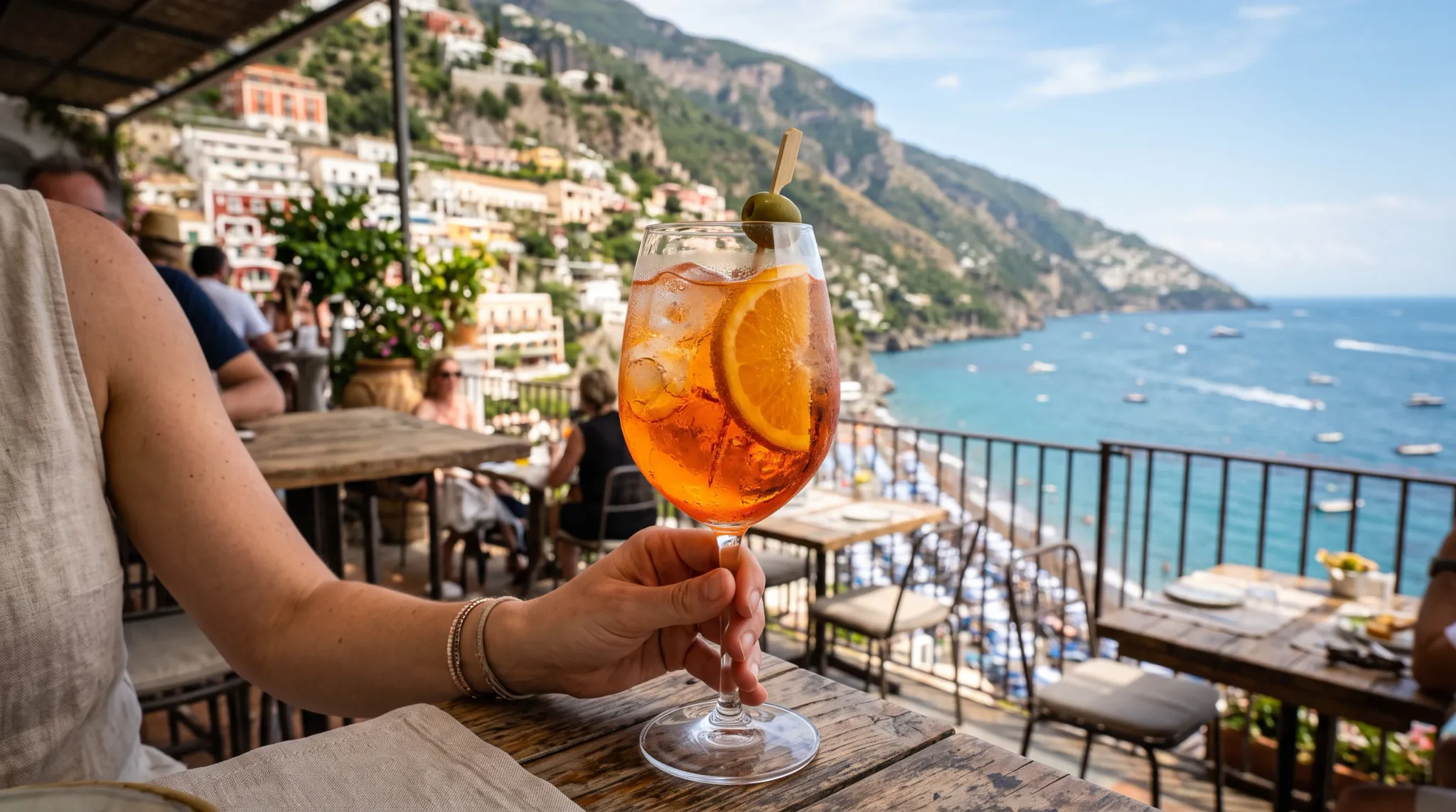 A seated foreground diner holds an orange iced aperol spritz garnished with an orange slice and green olive. The drink rests above a rustic wooden table on a sunny open-air terrace, with other diners, railings, colorful hillside buildings, steep cliffs, and a blue boat-dotted bay in the background.