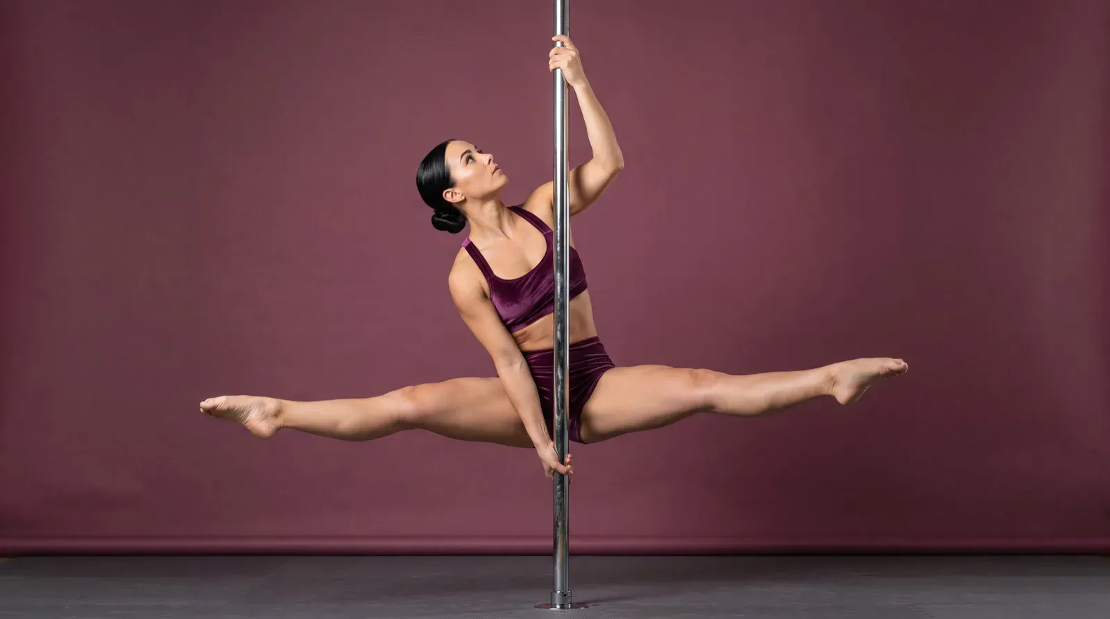 A woman in a purple velvet two-piece athletic outfit performs a full horizontal side split while suspended on a vertical metallic dance pole against a solid burgundy studio backdrop.