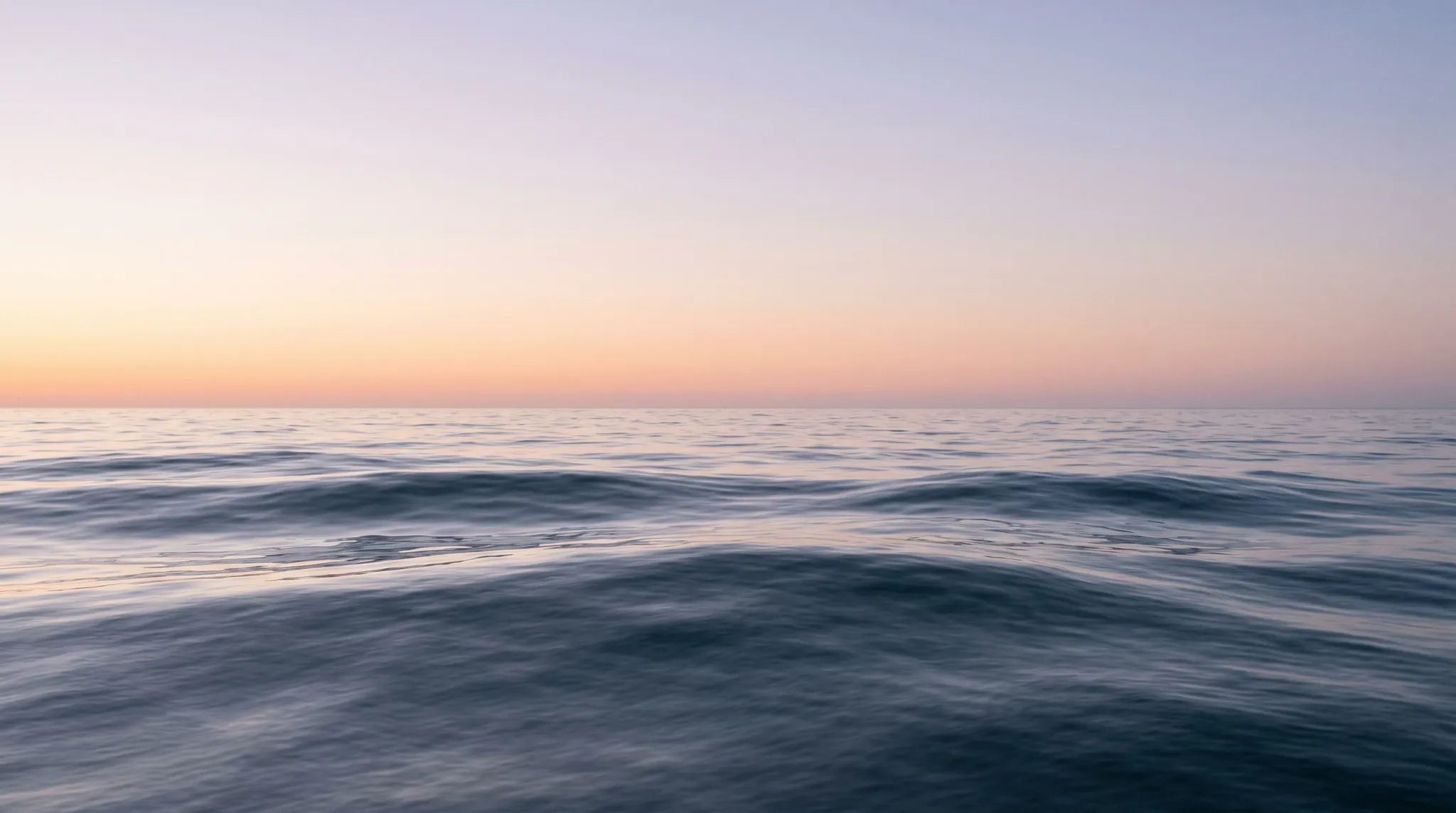 An expansive view of open ocean water with gentle rolling swells rippling across the foreground and midground, meeting a perfectly level horizon beneath a clear sky with a soft pastel gradient transitioning from peach to pale lavender at dusk.