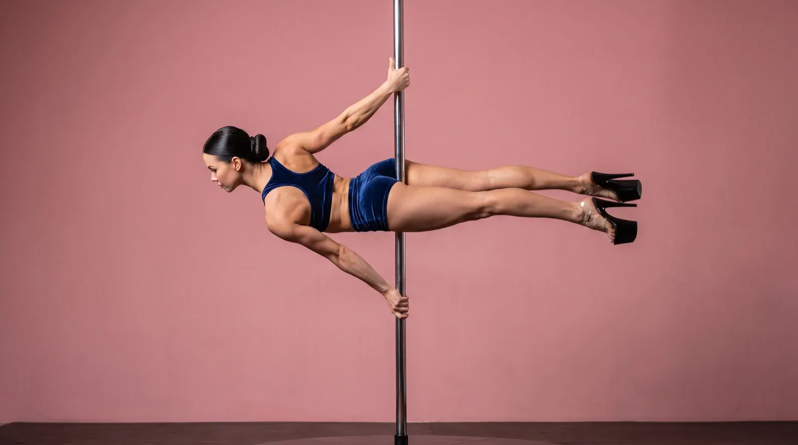 An athletic woman in a navy blue velvet two-piece outfit and black platform heels performs a horizontal flag hold suspended on a vertical metallic dance pole against a solid pink studio background.