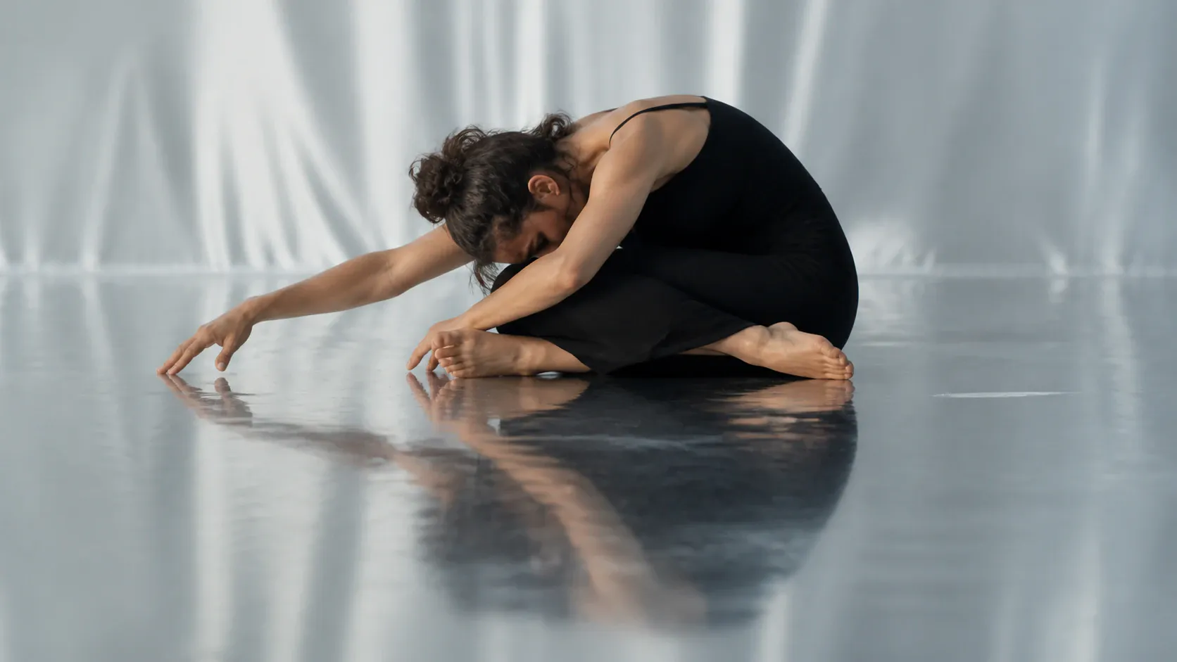A woman in a fitted black sleeveless dress-like garment sits cross-legged on a glossy studio floor, leaning forward with both arms extended and hands reaching toward the floor. Her dark curly hair is tied in a high bun, and a soft draped backdrop and clear floor reflection create a quiet minimalist studio scene.