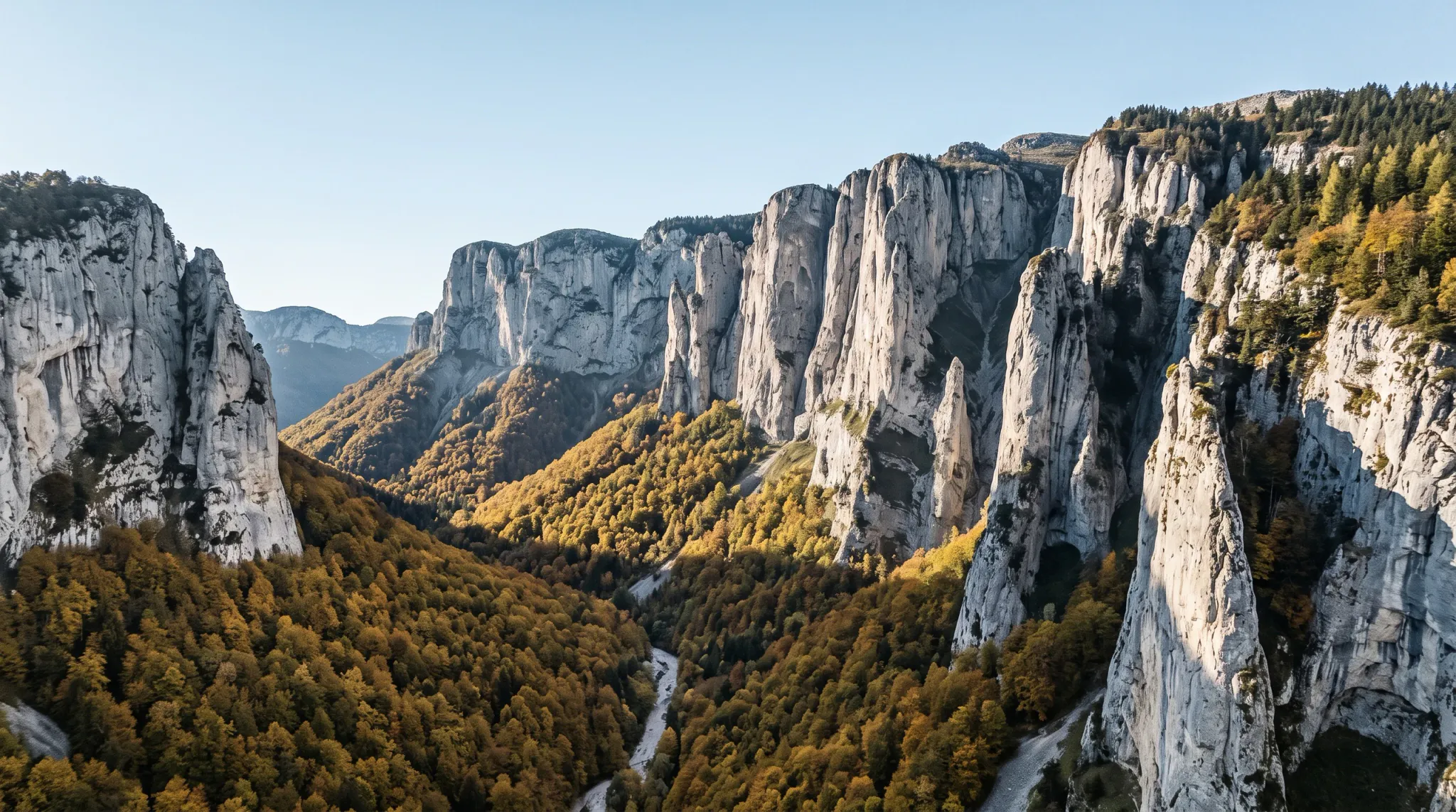 An aerial photograph captures a rugged mountain gorge flanked by sheer, sunlit limestone cliffs and towering rock spires. A dense forest displaying vibrant golden yellow, orange, and green autumn foliage carpets the valley floor, with a narrow stream winding through the canyon under a clear pale sky.
