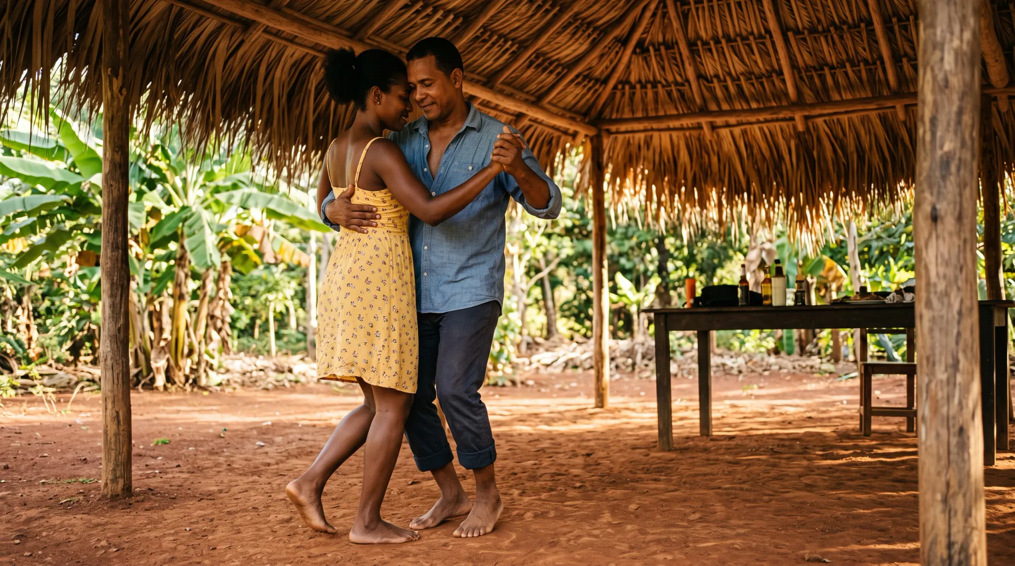 An adult man and woman dance intimately together barefoot on red soil beneath a rustic thatched-roof wooden shelter. The woman wears a yellow sleeveless sundress with a delicate floral print, while the man wears a light blue button-down shirt and rolled-up dark trousers. Surrounded by lush tropical greenery including banana plants, the couple smiles warmly as they hold hands and embrace in a peaceful rural setting.