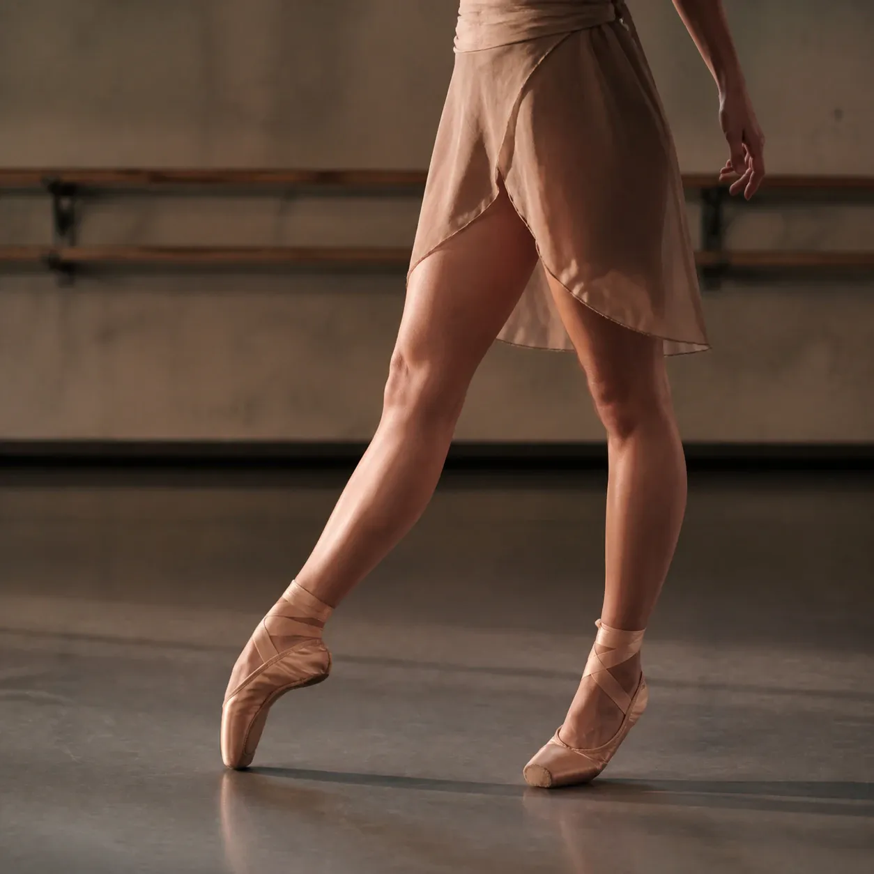 A cropped ballet dancer performs an extended pose en pointe on a warm studio floor, wearing a pale pink wrap skirt and matching satin pointe shoes. Wooden ballet barres line the softly lit background.
