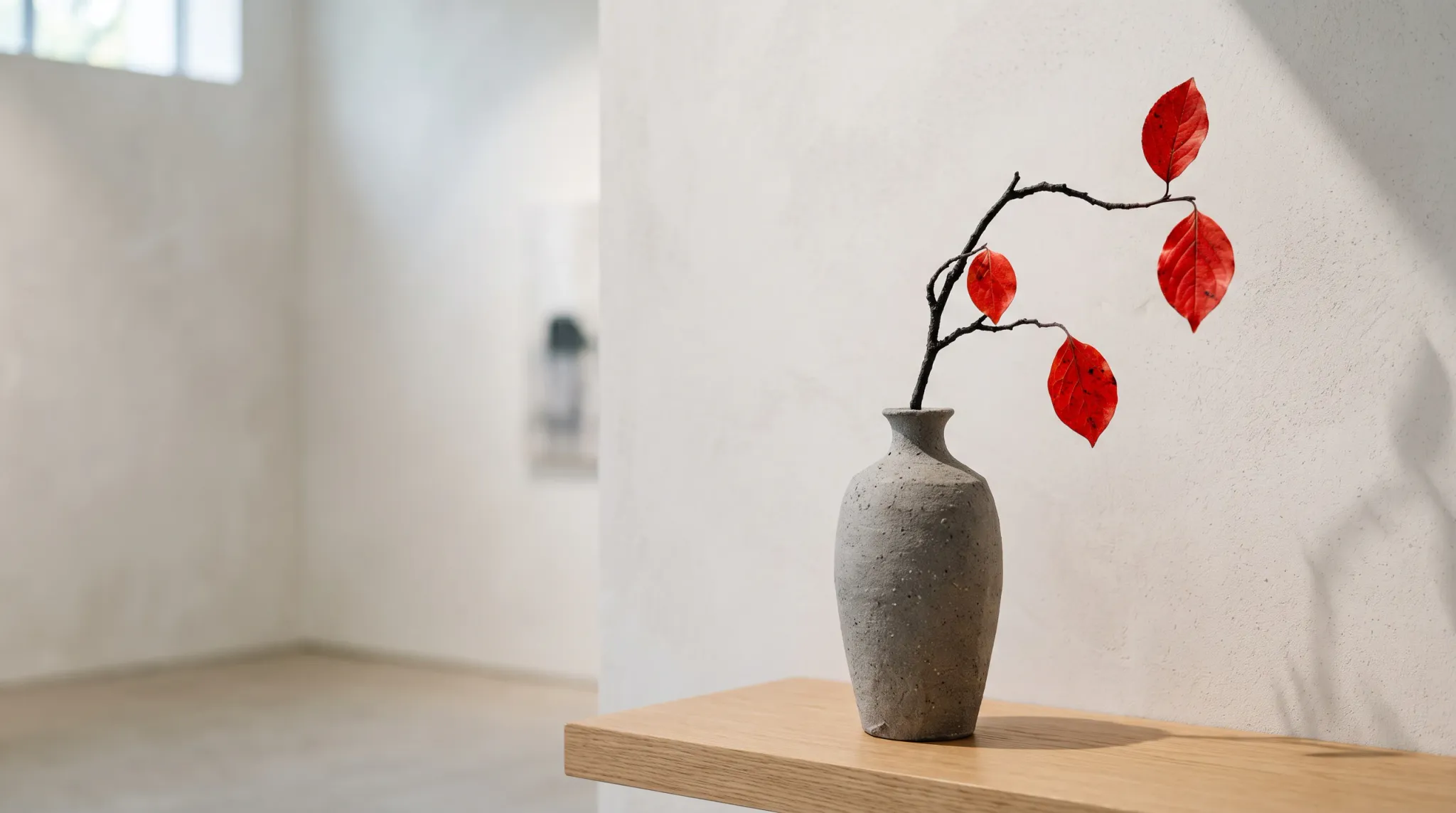 A minimalist still-life composition featuring a textured gray ceramic vase resting on a light-toned wooden shelf against a textured off-white wall. Inside the vase, a slender bare branch displays vibrant crimson autumn leaves. Soft natural daylight enters from an upper window on the left, casting subtle shadows across the wall and highlighting the tranquil, gallery-like interior.
