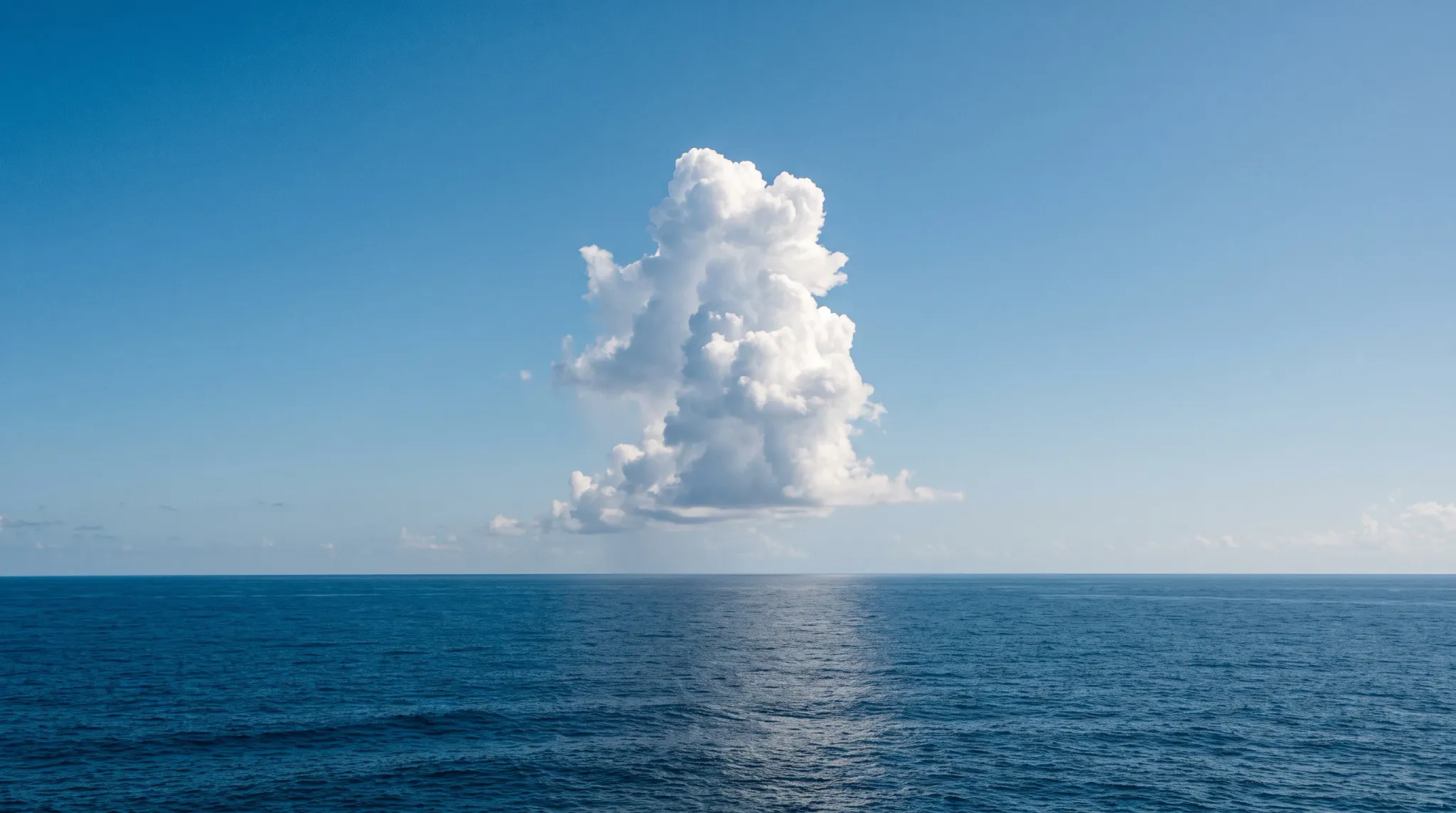 A solitary, vertical cumulus cloud formation rises prominently above a flat, level horizon over deep blue ocean waters under a clear azure sky in bright midday sunlight.