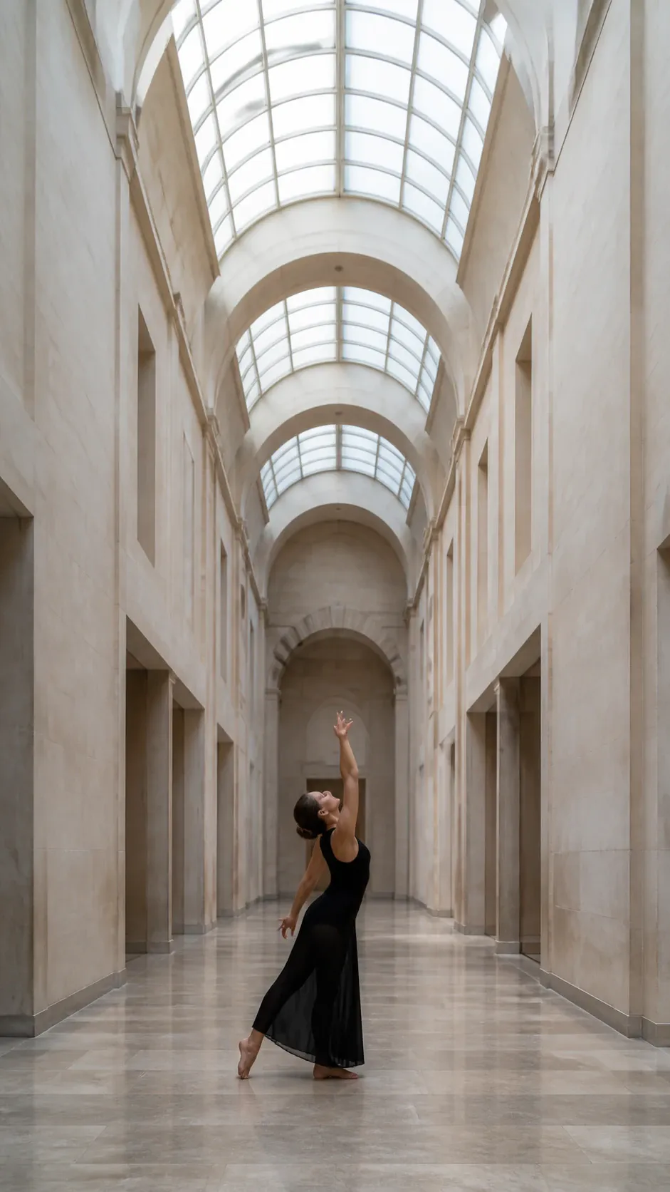 A full-length photograph of a female dancer in a black sleeveless dress striking an expressive pose with an arched back and raised arm inside a grand neoclassical hallway featuring towering stone arches, barrel-vaulted skylights, and polished stone flooring.