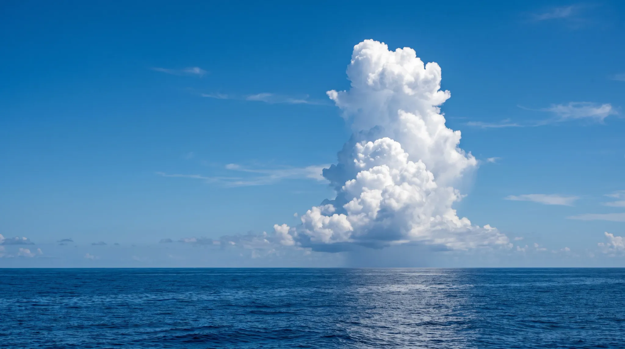 A wide landscape photograph of an expansive deep blue ocean under a clear daytime sky. A dramatic, towering vertical cumulus cloud formation rises near the center-right of the frame, with a visible localized rain shower falling from its dark flat base toward the rippled water surface below. The horizon is level and sharp across the lower third of the image, illuminated by bright natural sunlight reflecting on the sea.