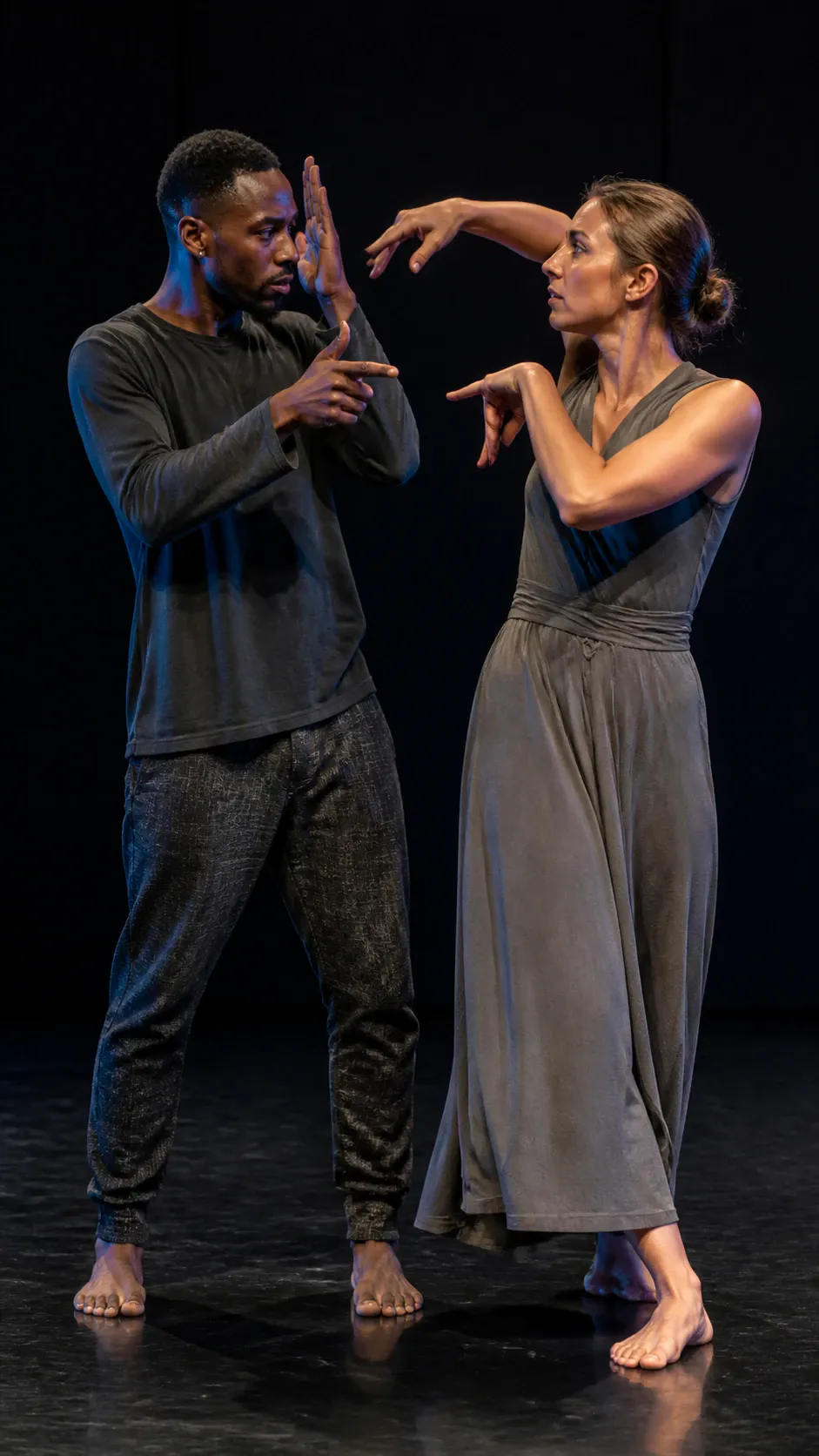 A full-length photograph of two barefoot contemporary dancers performing a choreographed duet on a dark stage. On the left, an adult male dancer wearing a charcoal long-sleeve top and textured pants gestures with raised hands toward his partner. On the right, an adult female dancer with her hair tied in a low bun wears a sleeveless gray wrap dress and mirrors the movement with bent arms and stylized hand shapes. The backdrop is dark and seamless under theatrical studio lighting.