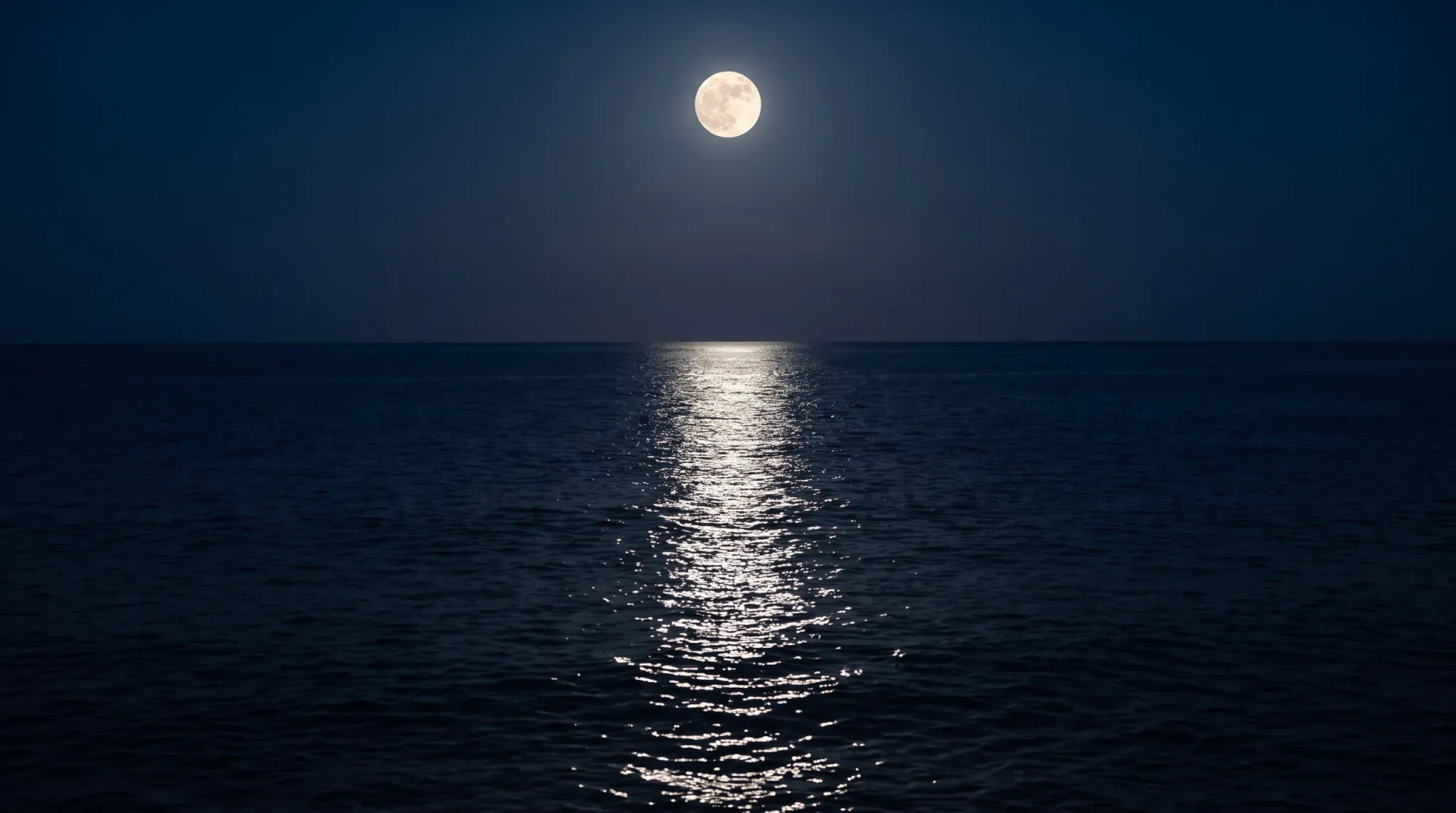 A glowing full moon hangs high in a dark blue night sky, casting a radiant moonlit reflection path across the surface of the calm ocean below toward the horizon.