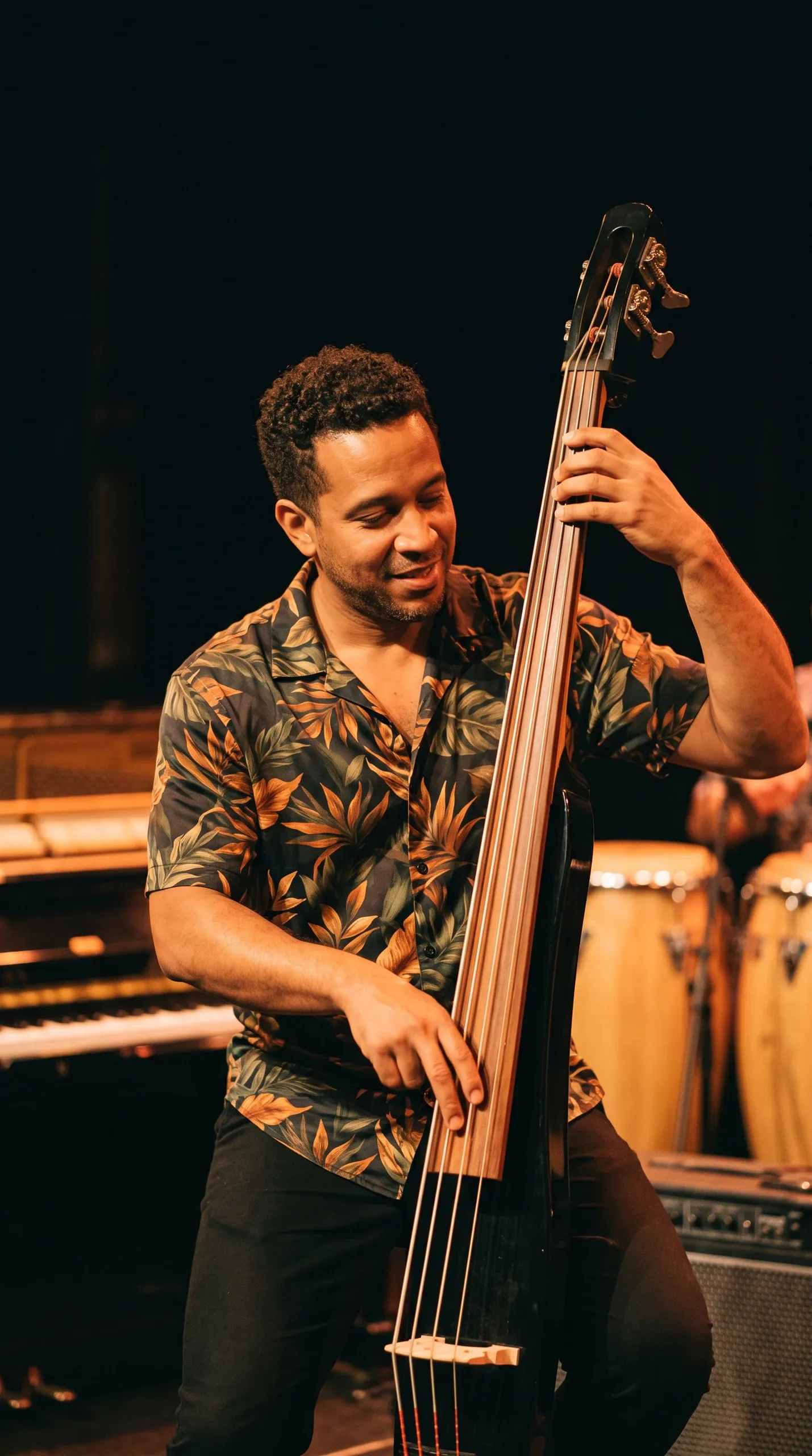 A smiling male musician in a dark floral short-sleeve shirt plays an electric upright bass on stage, accompanied by background instruments including conga drums and a piano in warm ambient stage lighting.