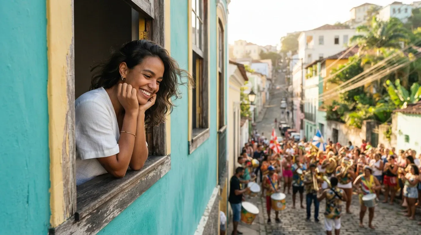 A smiling young woman in a white top leans out of a wooden window of a bright turquoise building with yellow trim, resting her face on her hands while watching a colorful street carnival parade and drum band on a cobblestone road below.