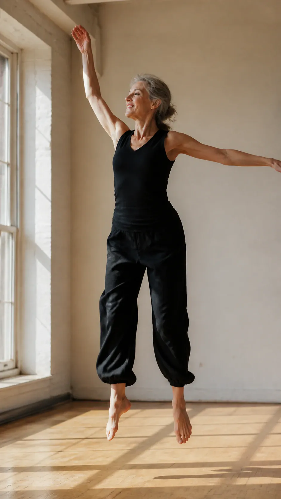 A full-body shot of a smiling mature woman with gray hair leaping barefoot into the air inside a bright, sunlit dance studio with hardwood flooring. She wears a sleeveless black tank top and loose cropped black trousers, raising one arm high and extending the other as natural daylight streams through a large window.