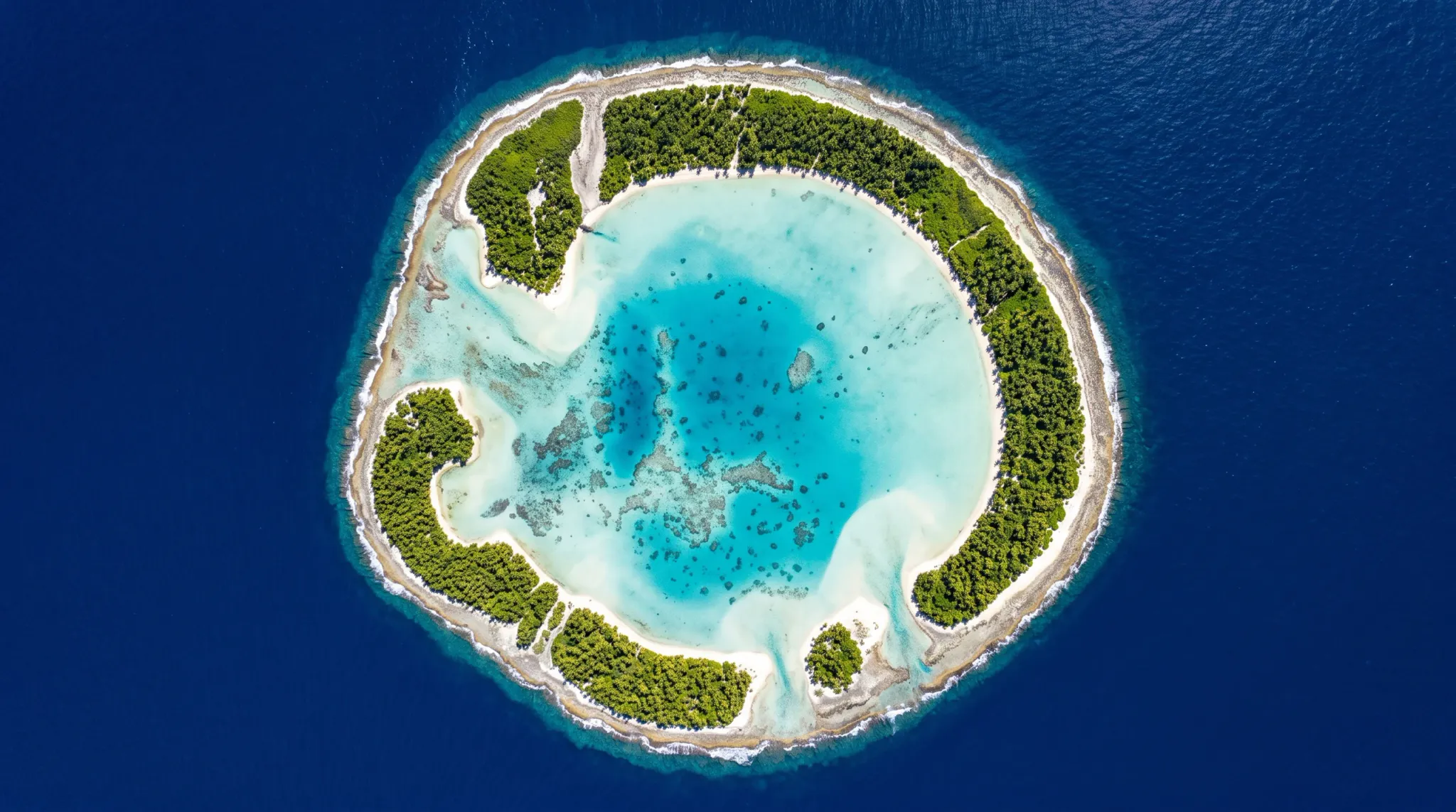 A high-angle top-down aerial photograph of a circular tropical coral atoll surrounded by deep blue ocean waters. The ring of the atoll is lined with lush green palm trees and white sand beaches, enclosing a central turquoise lagoon with visible submerged coral reefs and sandbars. Bright sunlight directly illuminates the calm clear shallow water, highlighting vivid contrasts between the turquoise lagoon and deep navy sea.