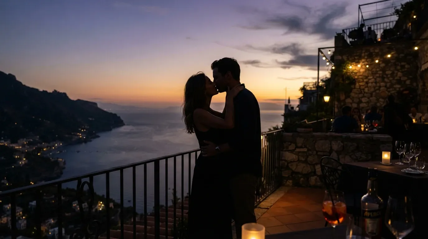 A romantic silhouette of a man and woman embracing and kissing on a restaurant balcony overlooking the sea at sunset. An Aperol Spritz bottle and glasses sit on a candlelit table in the foreground, with other diners enjoying the view in the background under string lights. The dramatic coastal mountains feature twinkling town lights in the distance.