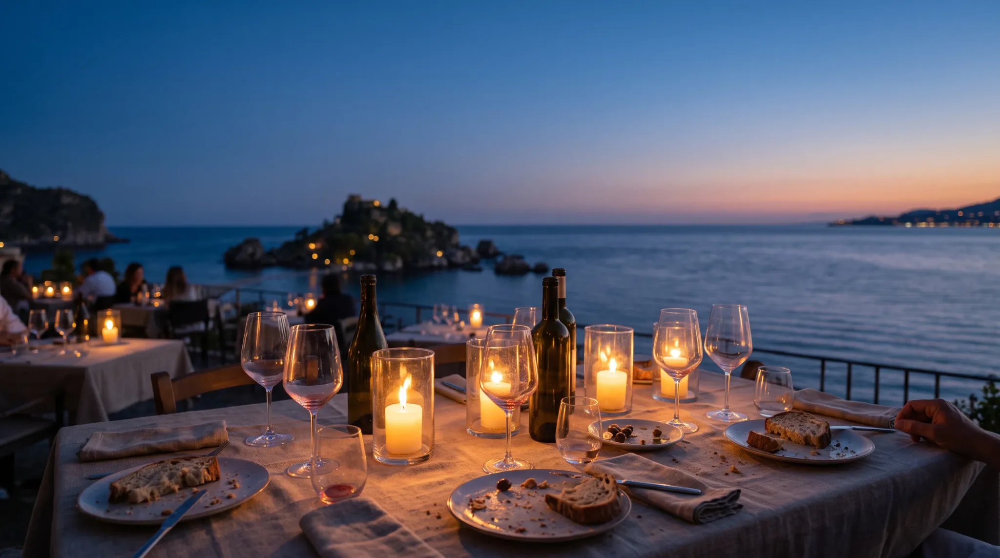 An elegant outdoor dining table with wine, bread, candles, glasses, plates, cutlery, and linen napkins overlooks a calm, rippled sea and a small rocky island at sunset. Several blurred diners occupy tables in the background, with distant coastline lights visible across the water.