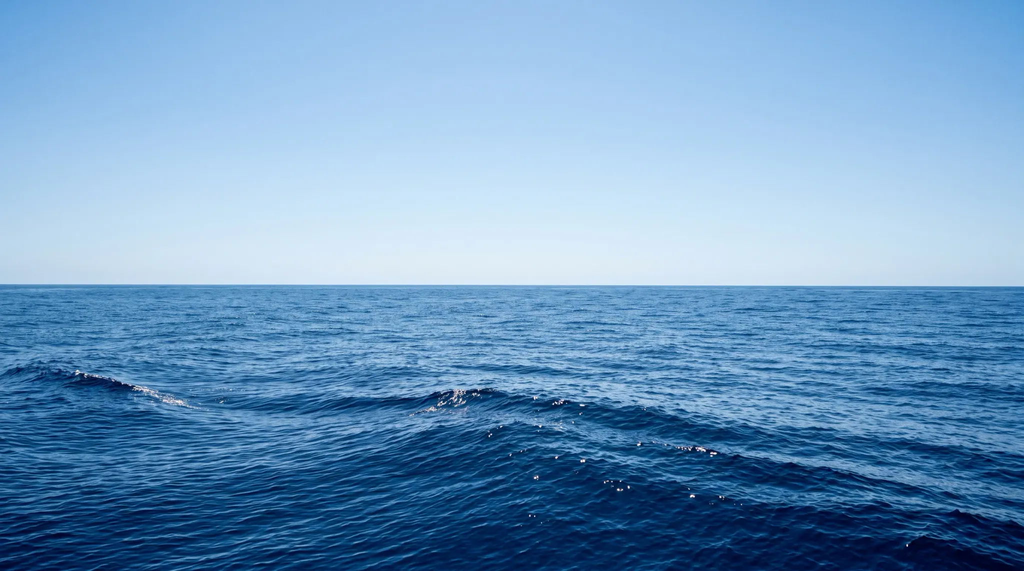 Wide, minimal seascape showing deep blue rolling ocean waves beneath a bright clear blue sky, with a level horizon near the center and no land, boats, or people visible.