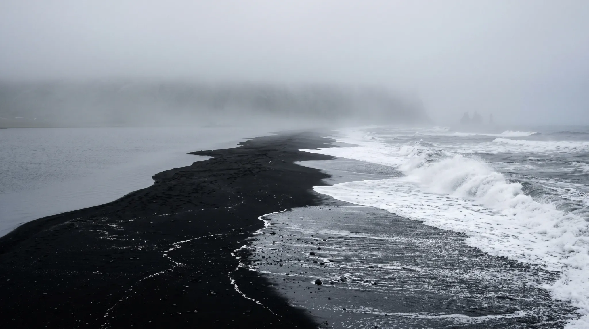 A wide coastal landscape shows a dark black-sand shoreline narrowing into dense fog, with calm water on the left and powerful white waves breaking along the right. Distant sea stacks are faintly visible through the mist beneath a pale gray sky.