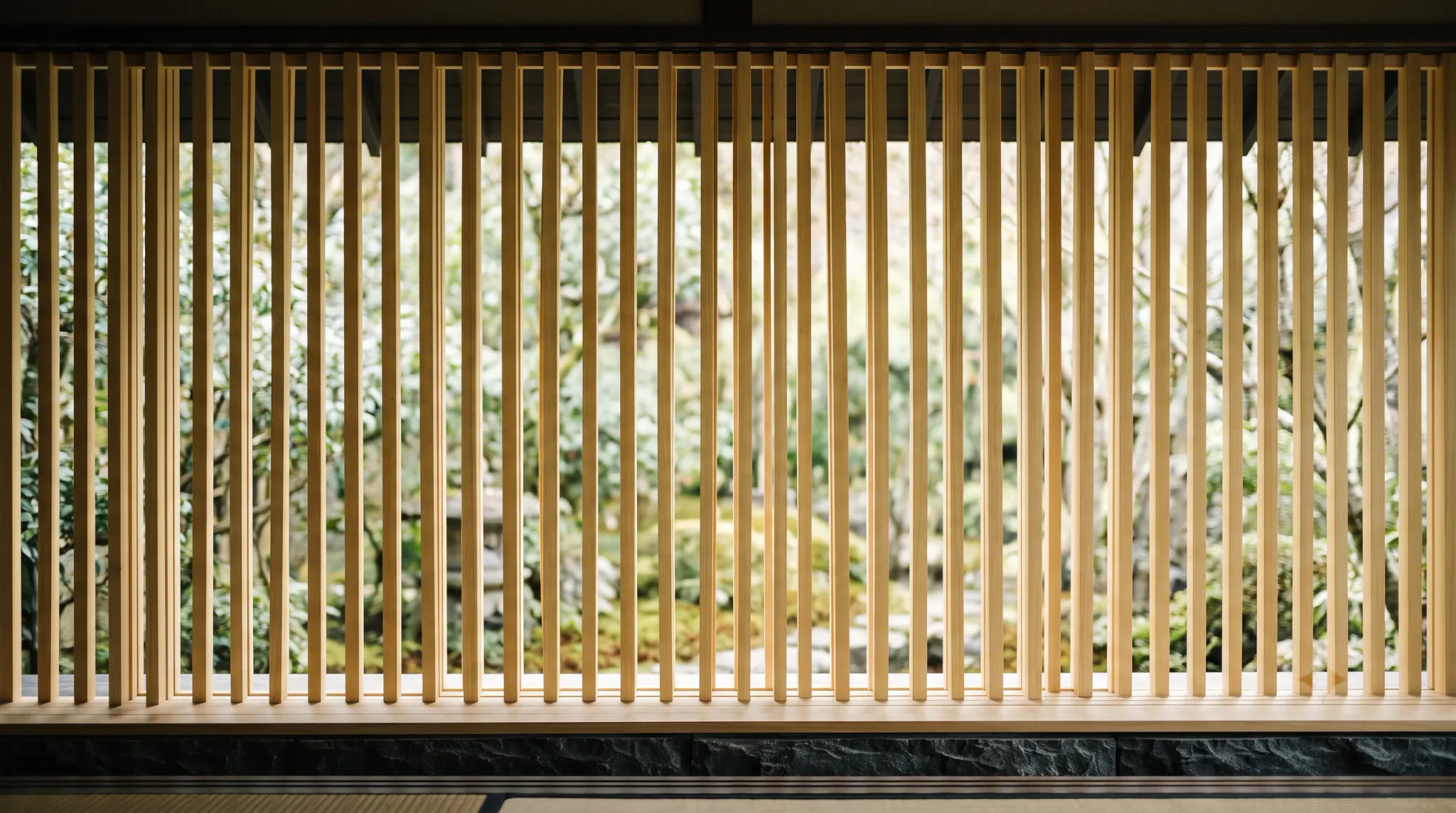 A symmetrical eye-level interior view looking through vertical light-wood lattice slats toward a lush Japanese garden softly blurred in the background. The foreground shows a smooth wooden sill and dark textured stone ledge above a tatami floor, contrasting with the vibrant green foliage outside.