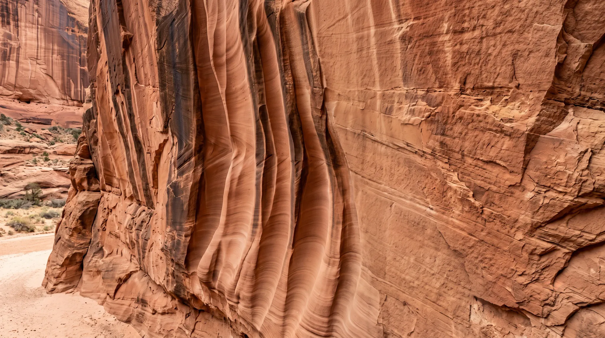 A wide color photograph shows a massive warm red-orange sandstone canyon wall with dramatic vertical fluted grooves, smooth wind- or water-sculpted curves, horizontal strata, cracks, and rough weathered surfaces. A sandy rocky canyon floor and sparse shrubs appear at the left edge and in the distance; no people, buildings, or vehicles are visible.