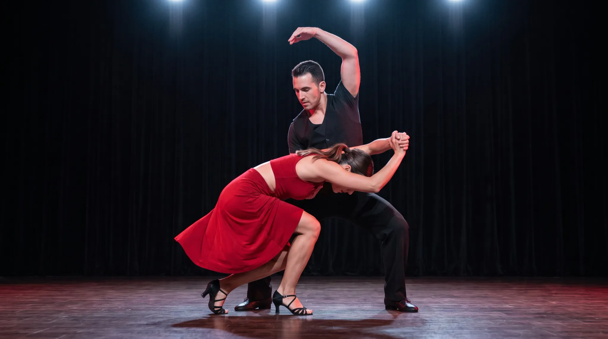 Two adult dancers perform a dramatic partnered dance pose on a theater stage. A woman in a flowing red sleeveless dress crouches and leans forward beneath a man in black, who bends behind her with one arm arched overhead. They hold hands while illuminated against black curtains and a polished wooden floor.