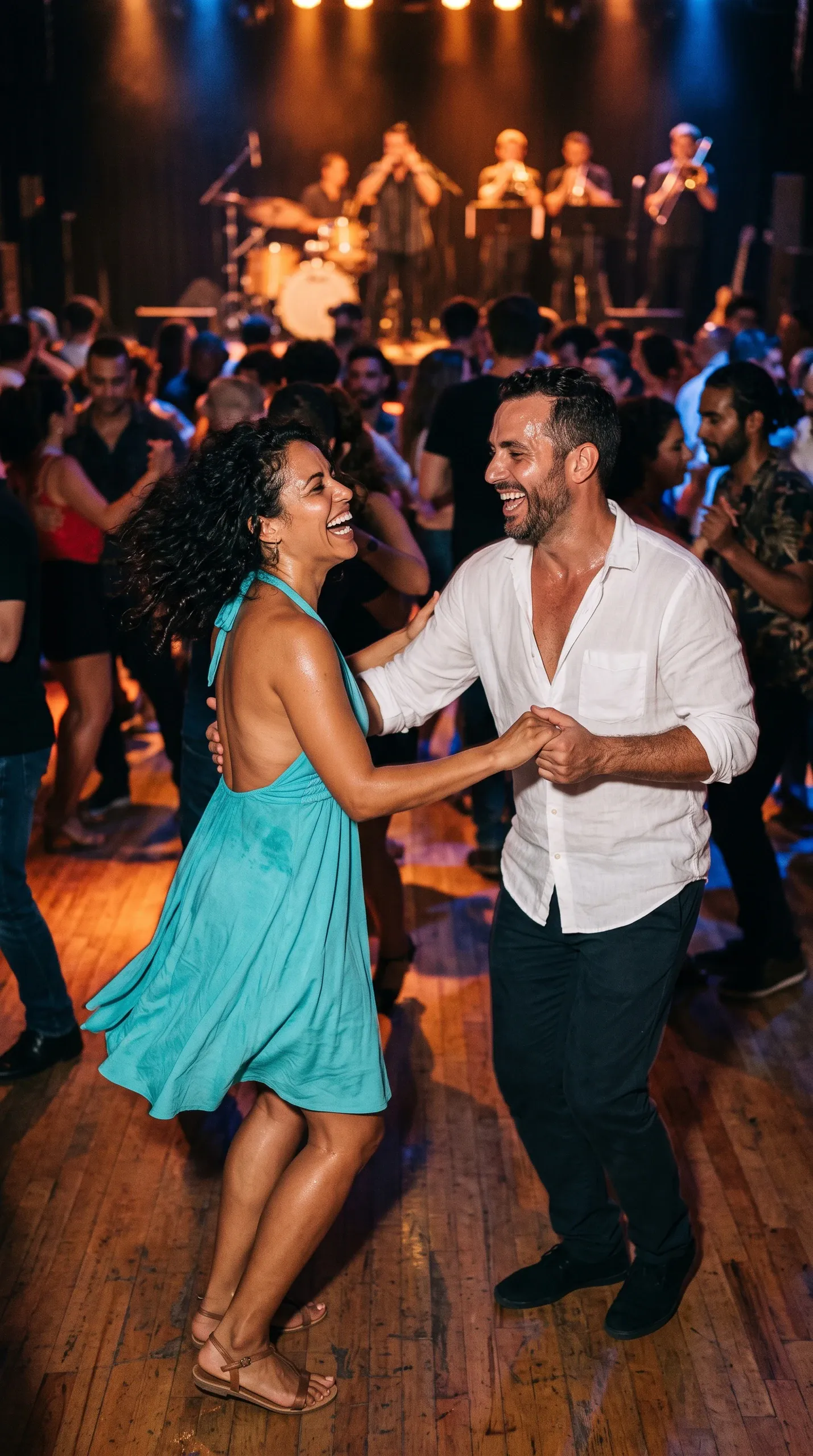 A smiling woman in a vibrant turquoise halter-neck dress and a man in a white button-down shirt and dark trousers dance together holding hands on a polished wooden floor in a lively, crowded nightclub. In the background, a live brass band performs under warm stage lights while other patrons dance.