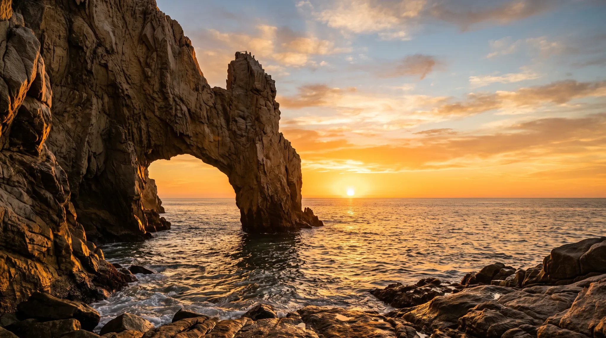 A wide-angle landscape photograph capturing a towering natural rock arch standing in the ocean during sunset, with the golden sun low on the horizon casting warm light across the rugged rock faces and shimmering water.