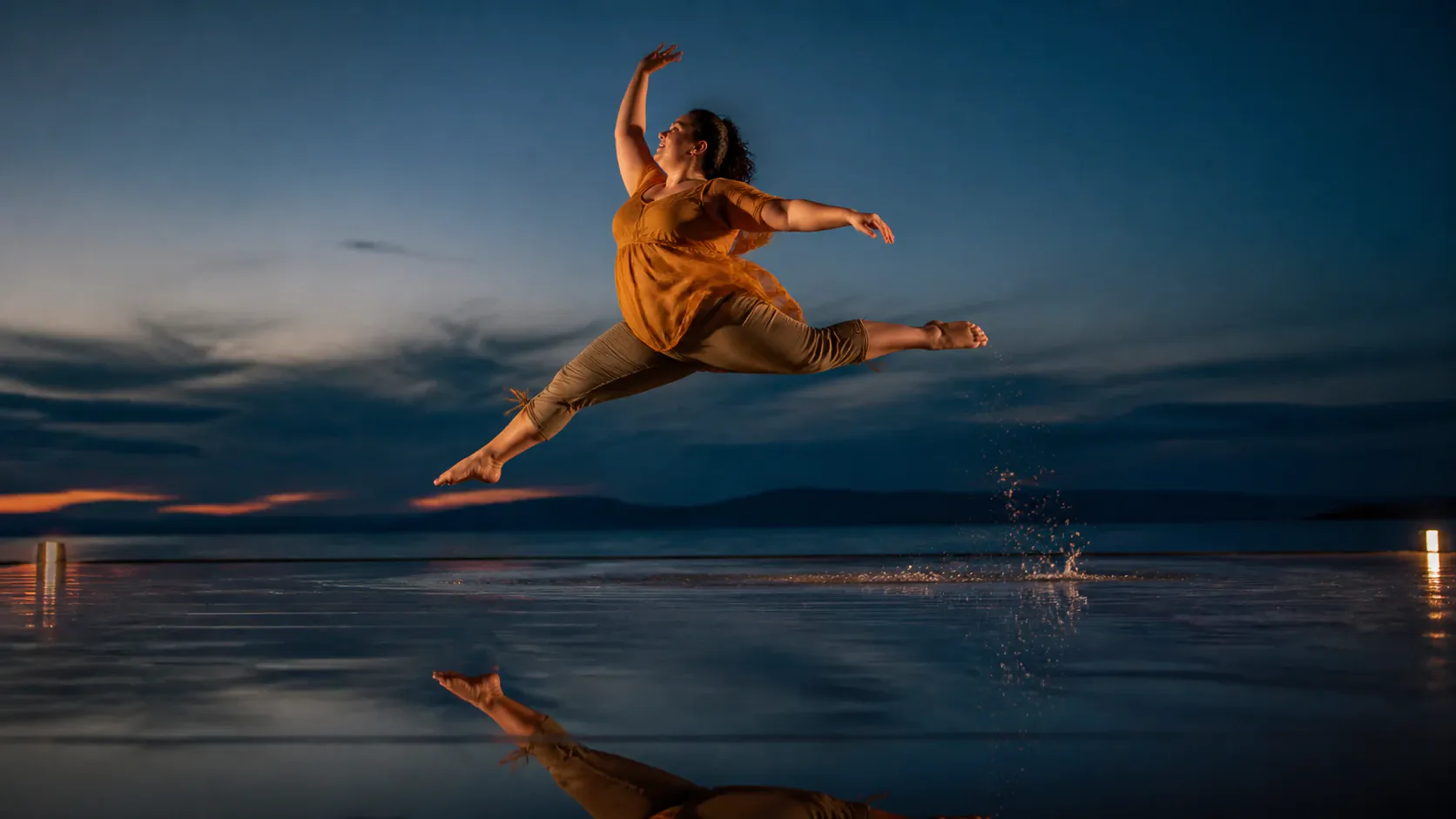 A female dancer leaps dynamically mid-air in a grand jeté split leap over a shallow, reflective wet surface at dusk. She is dressed in a short-sleeved orange top and cropped beige trousers, barefoot with arms outstretched and head lifted in a joyful expression. The mirror-like surface below reflects her airborne form along with splashing water droplets against a dark twilight sky and a distant horizon glowing with the last light of sunset.