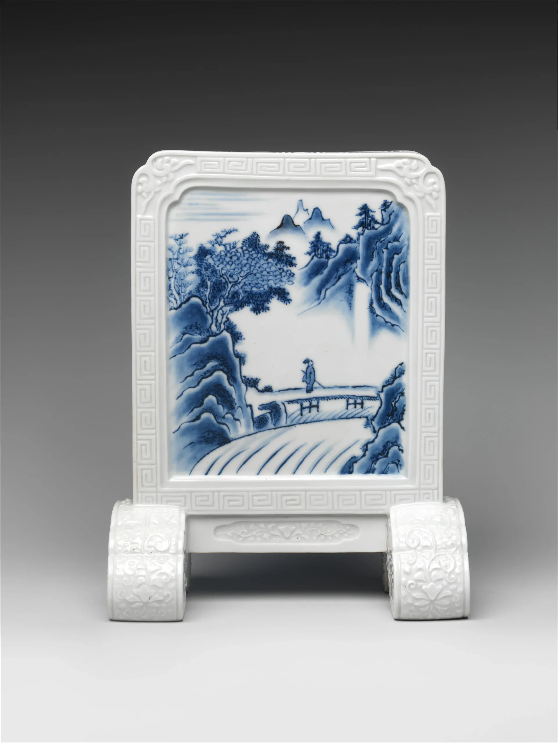 A glossy white ceramic chair or decorative stand is centered against a gray studio backdrop. Its rectangular backrest has a blue-and-white mountainous landscape scene, with raised floral scrollwork and geometric key-pattern borders on the frame and supports.