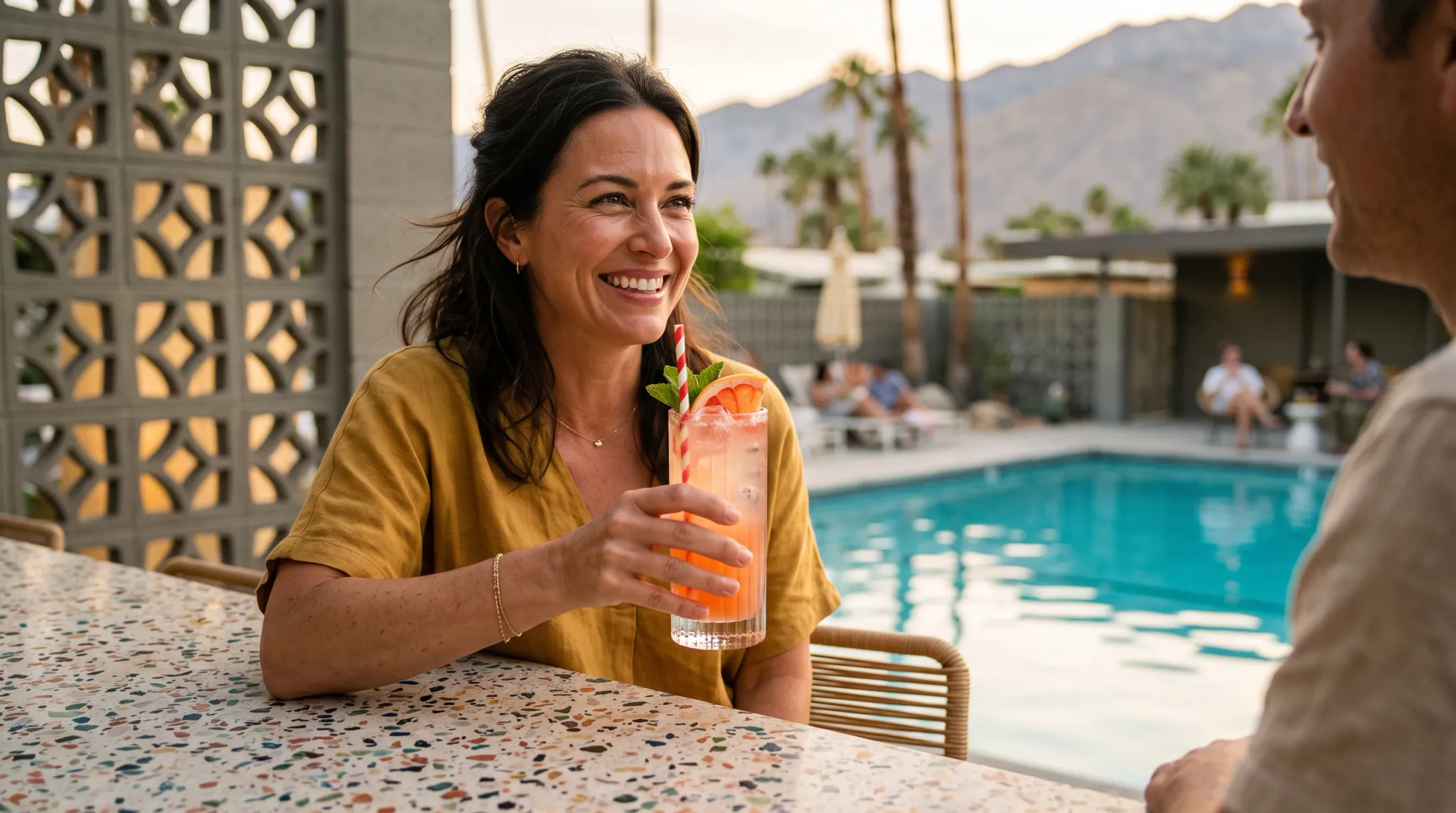 A smiling adult woman with dark brown, loosely wavy, shoulder-length hair sits at a terrazzo-topped outdoor bar counter beside a turquoise swimming pool. She wears a mustard yellow short-sleeve linen-looking top and holds a tall glass containing a pink/orange cocktail garnished with a grapefruit slice, fresh mint and a red-and-white striped straw. A thin gold necklace and a delicate gold bracelet are visible. She laughs and faces slightly right toward an out-of-frame companion, whose shoulder and back of head appear at the far right edge of the frame. In the background, palm trees, a mountain range, and a mid-century modern building with decorative concrete breeze-block screen wall are visible in warm golden-hour light. Several people relax in blurred background near the pool. A folded beach umbrella stands poolside.