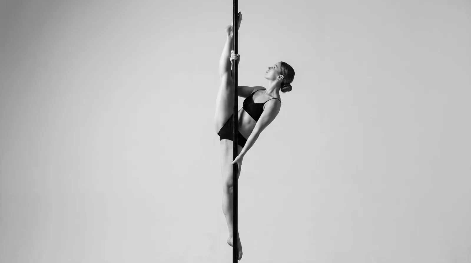 A monochrome studio photograph showing a female dancer suspended on a vertical pole while performing a full vertical split. She wears a fitted black sports bra and matching shorts, gripping the pole with both hands as she gazes upward against a seamless, well-lit studio background.