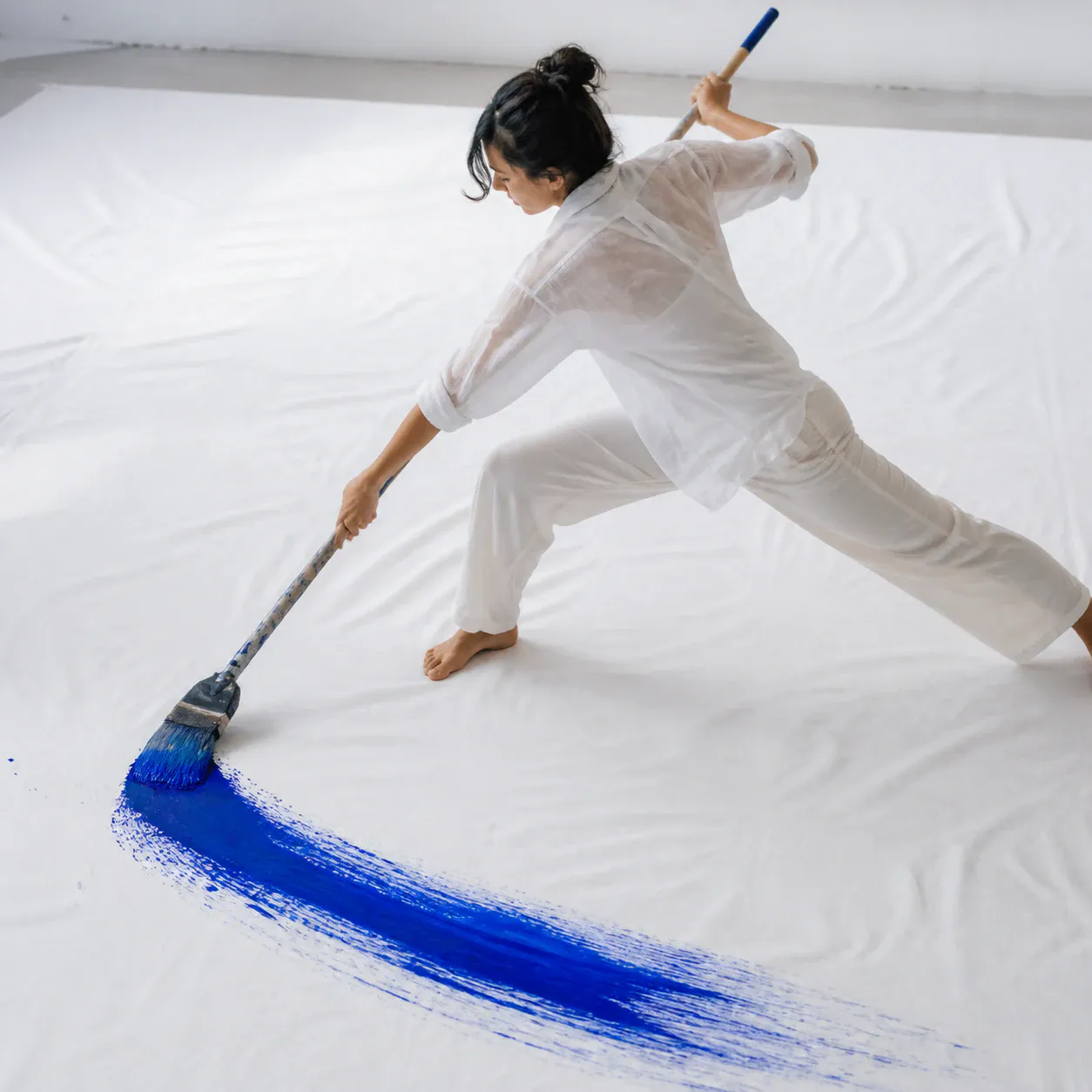 A single adult person in loose white clothing lunges across a bright, minimal indoor studio while painting a broad vivid-blue stroke across a large white canvas or floor covering. The person is barefoot, leaning forward with both arms extended, and holds a long paintbrush.