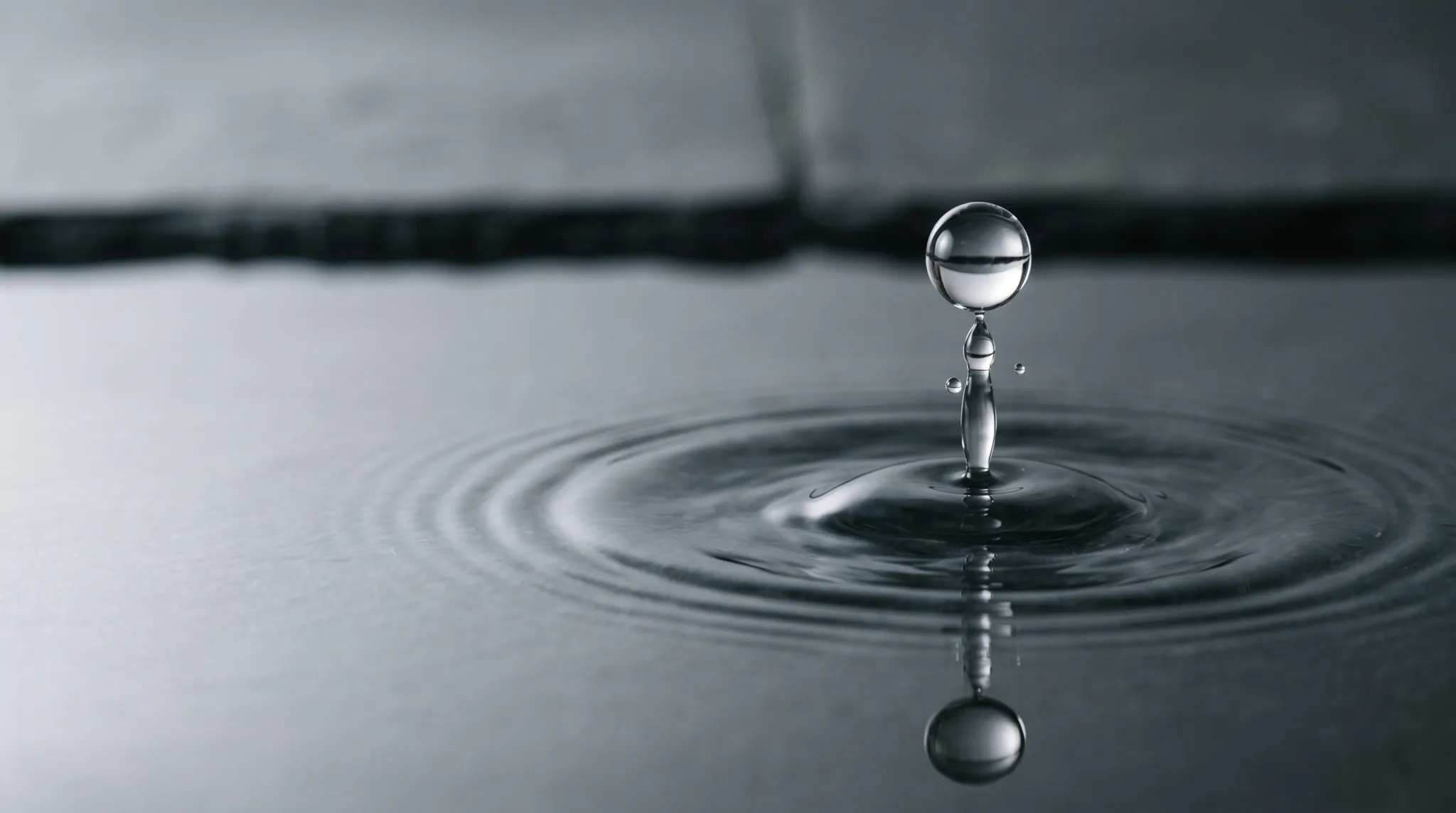 A spherical water droplet rises above a dark, calm water surface while a narrow splash column connects it to the impact point. Concentric ripples spread outward, with a mirrored droplet reflection below. The image is monochrome, softly lit, minimally composed, and uses shallow depth of field.