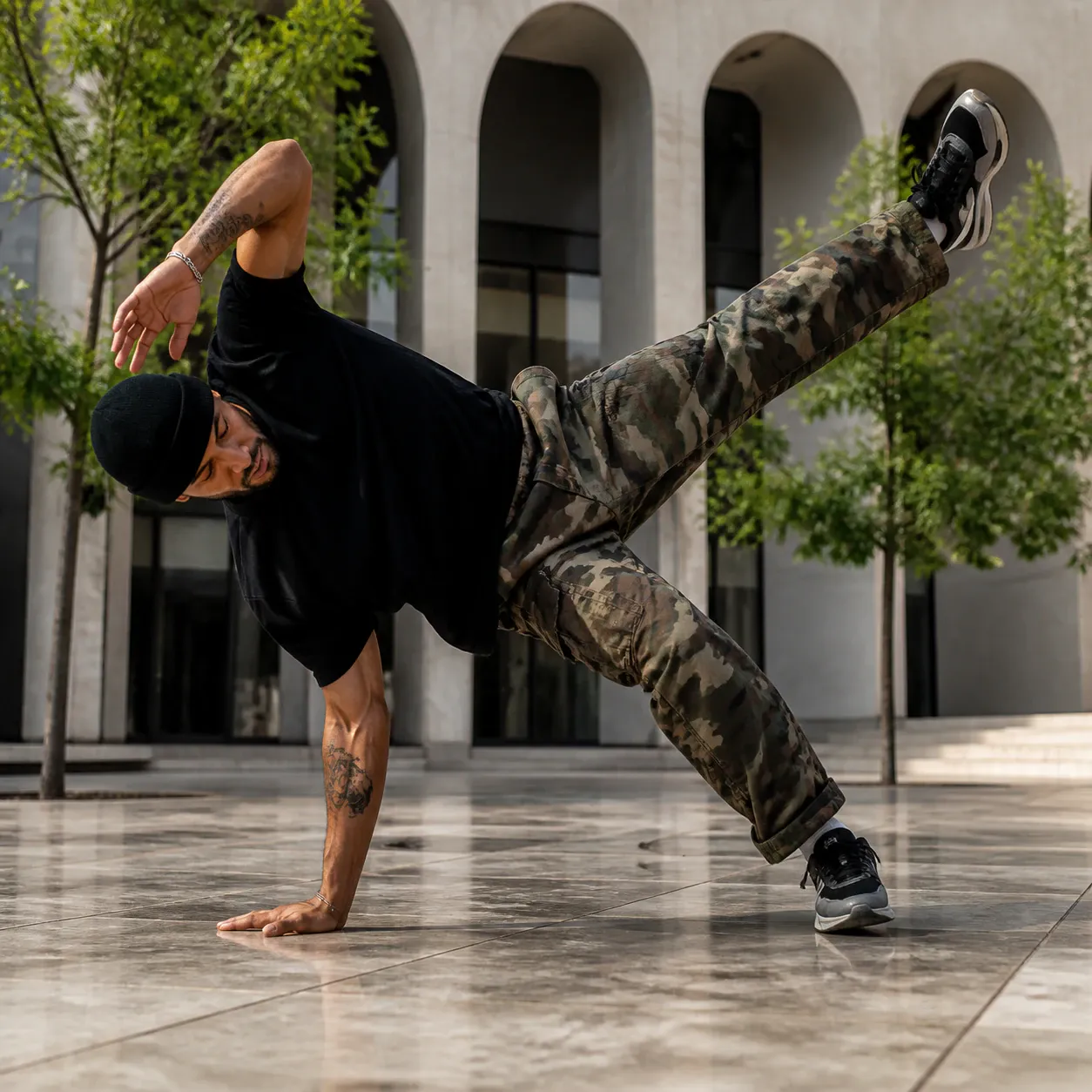 A young athletic male breakdancer balances dynamically on one hand and one foot in an outdoor stone plaza. He wears a black beanie, black t-shirt, camouflage cargo trousers, and black sneakers, with visible tattoos on his forearms. Behind him stands modern architecture featuring monumental stone arches and slender green trees.