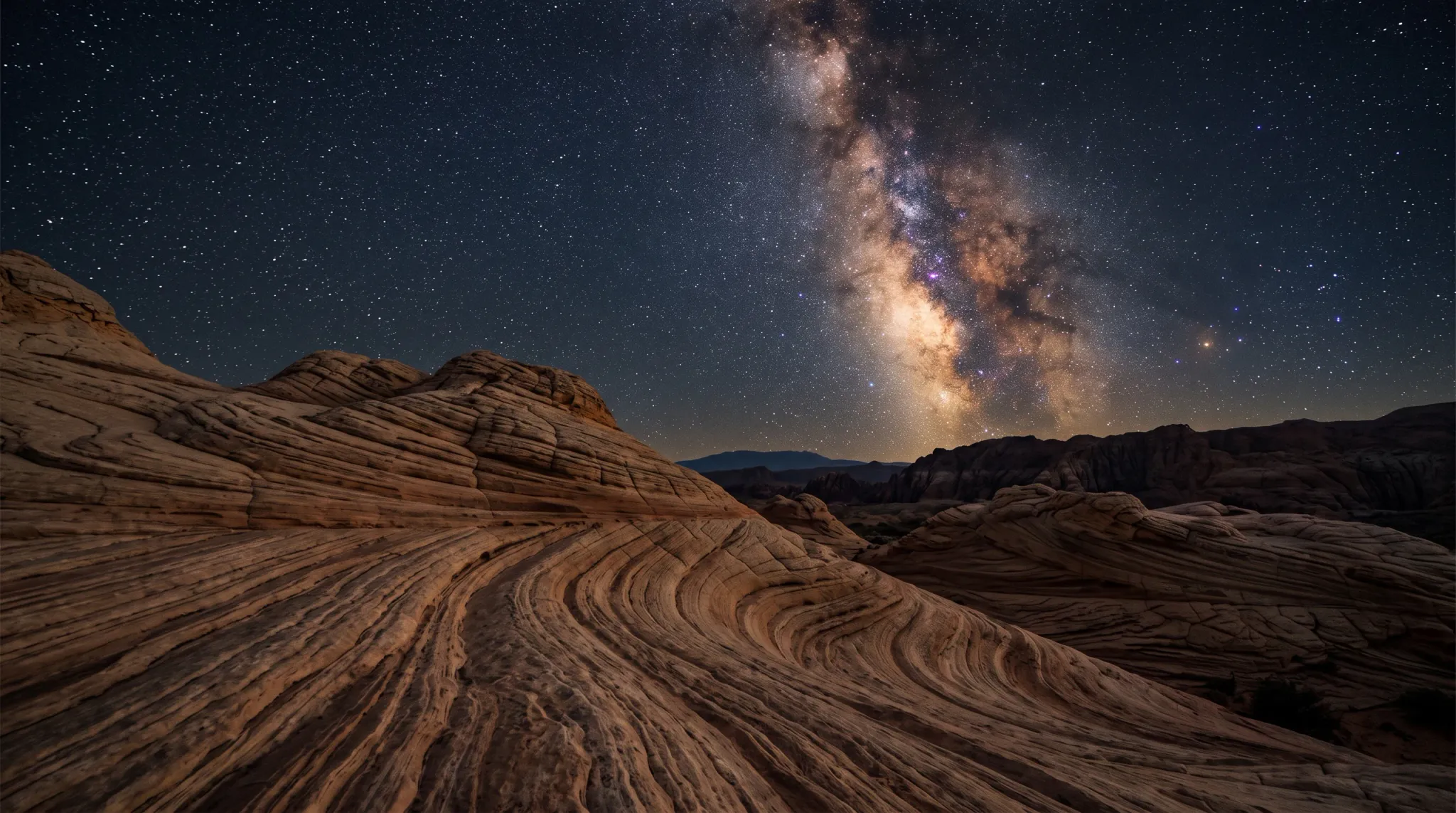 A wide nighttime landscape view showing prominent, undulating sandstone rock formations in the foreground under a vast starry sky. The Milky Way is clearly visible as a bright band stretching across the dark, clear sky. In the distance, dark silhouettes of a mountain range line the horizon.