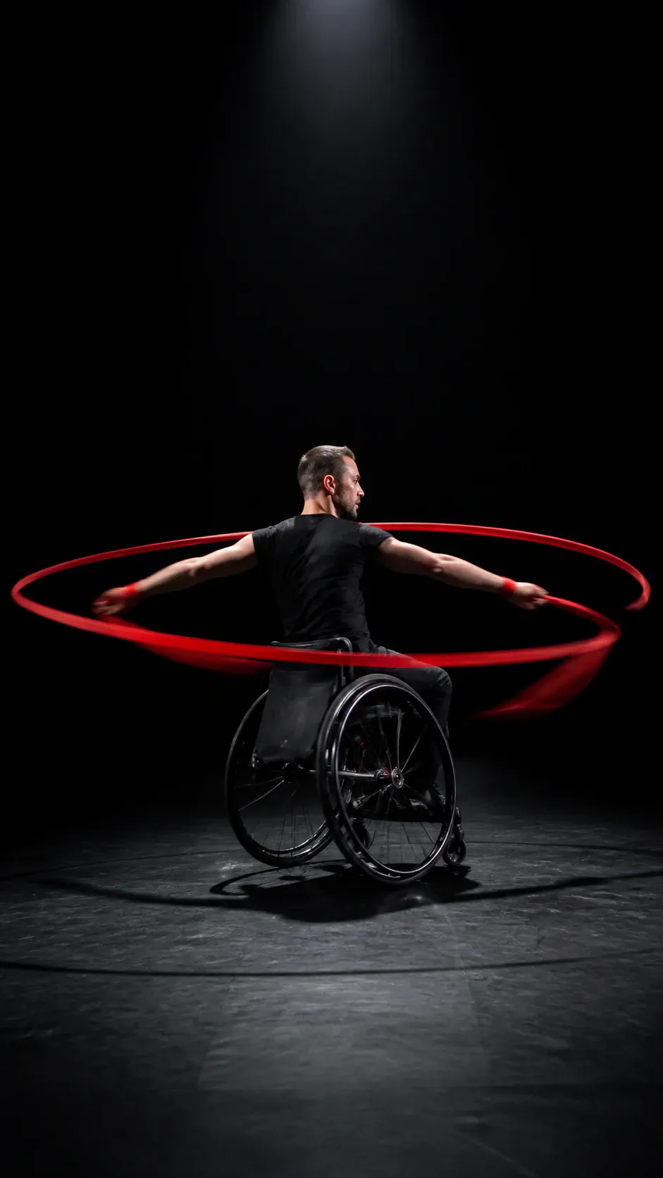 A single adult-looking man in a black T-shirt sits in a black wheelchair on a studio floor, facing mostly away while turning his head to the right. His arms extend sideways and his hands grip a bright red hoop that appears to be spinning around him, creating strong motion blur. Dramatic overhead studio lighting isolates the performer against a nearly black background.