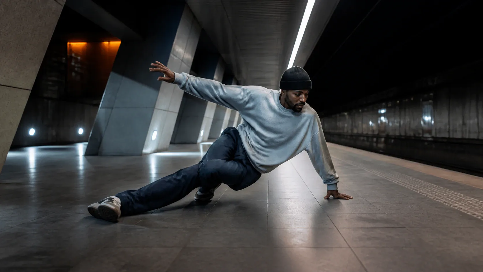 A single adult man in a black beanie, gray sweatshirt, dark trousers, and sneakers performs a low breakdance-style floor move on a tiled subway station platform. One hand supports him on the floor while his other arm extends outward; his legs are spread with one bent and one extended. Overhead strip lighting illuminates the spacious, dark modern station interior.