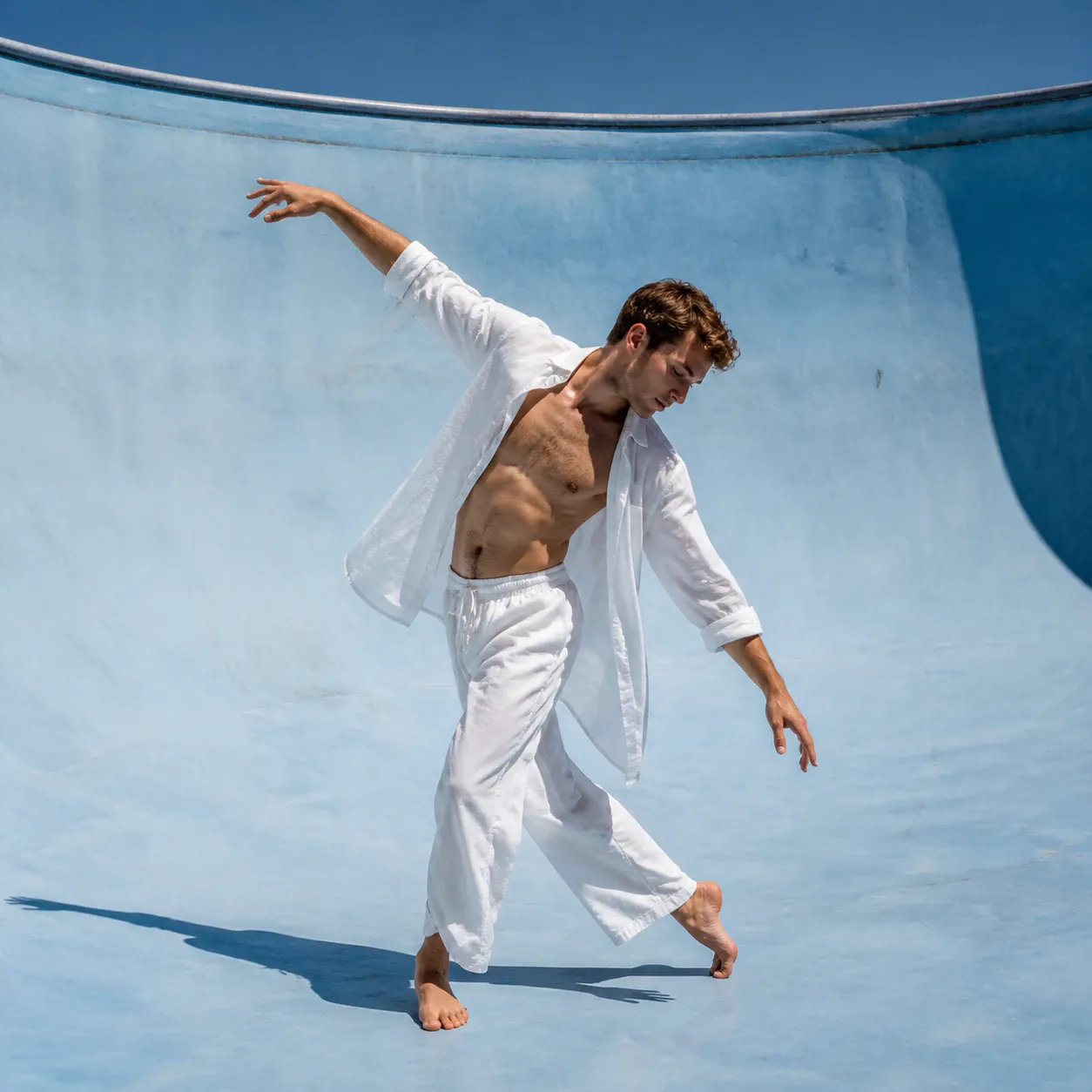 A full-body shot of a young adult male dancer posing gracefully inside an empty curved light-blue pool basin. He is barefoot, wearing an unbuttoned white long-sleeved shirt that reveals his torso and loose white ankle-length trousers. Both arms are outstretched in an elegant diagonal dance line under bright direct sunlight.