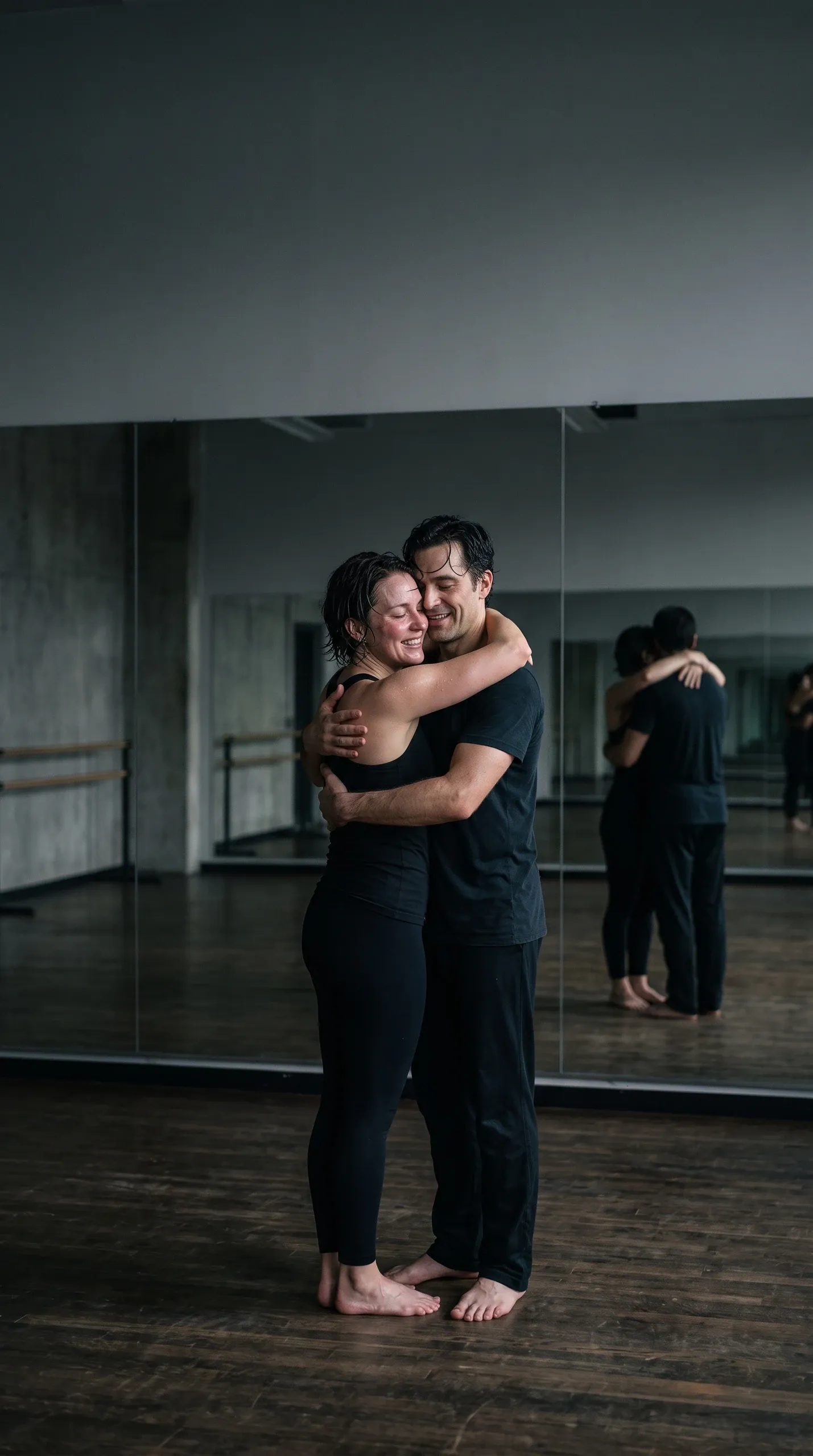 A female and male contemporary dancer share a warm, affectionate embrace in an empty rehearsal studio. Both are barefoot, dressed in black athletic dancewear, and visibly glistening with sweat from an intense practice session. The woman rests her head against her partner smiling with eyes closed, while the man smiles holding her close. Behind them, large wall mirrors reflect their embrace across the polished wooden dance studio floor.